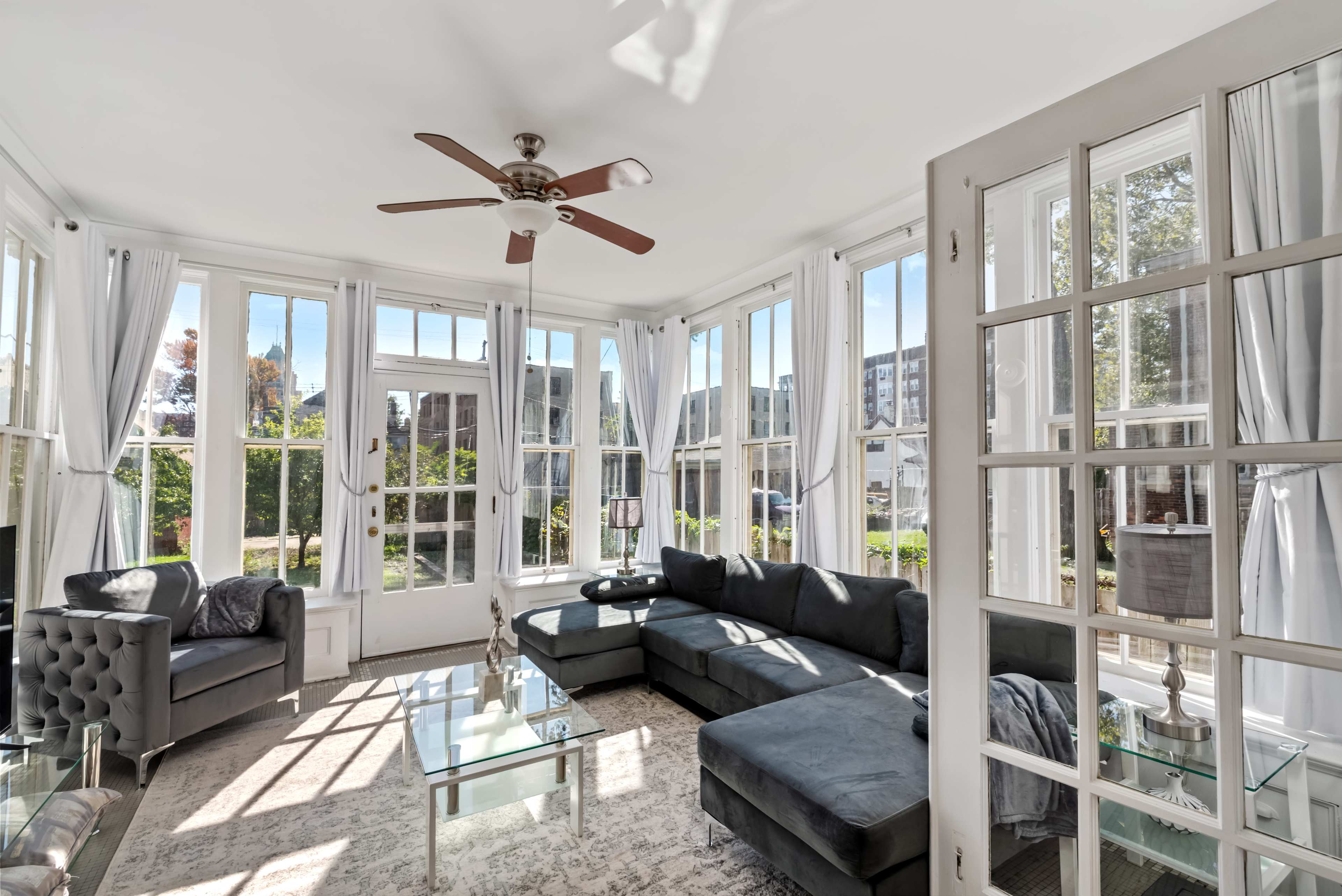 A sunlit sunroom features large windows, a sectional sofa, a glass coffee table, and a ceiling fan.