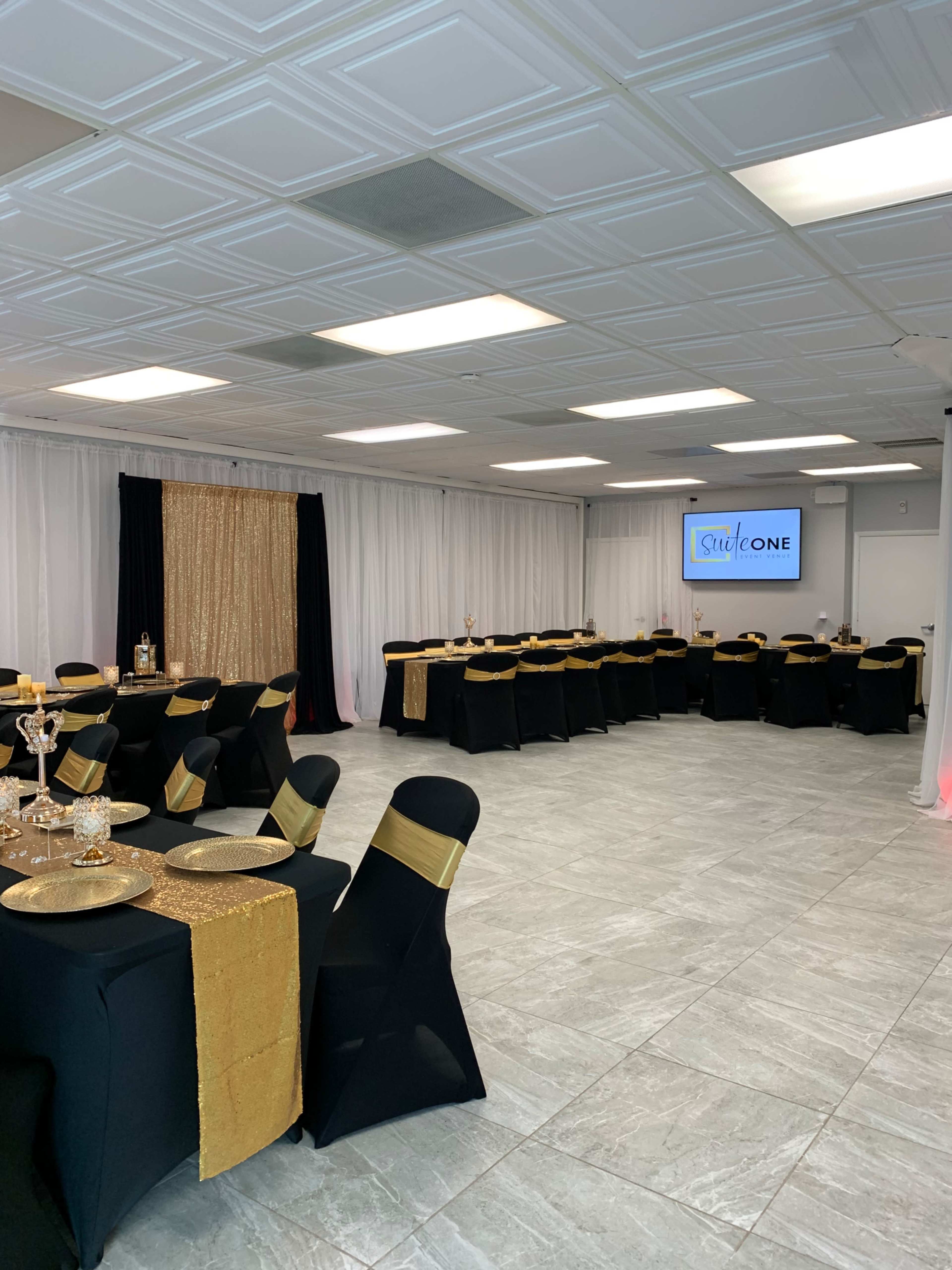 A banquet hall set up with black tablecloths adorned with gold accents, featuring tables arranged for an event and a large screen displaying a logo.