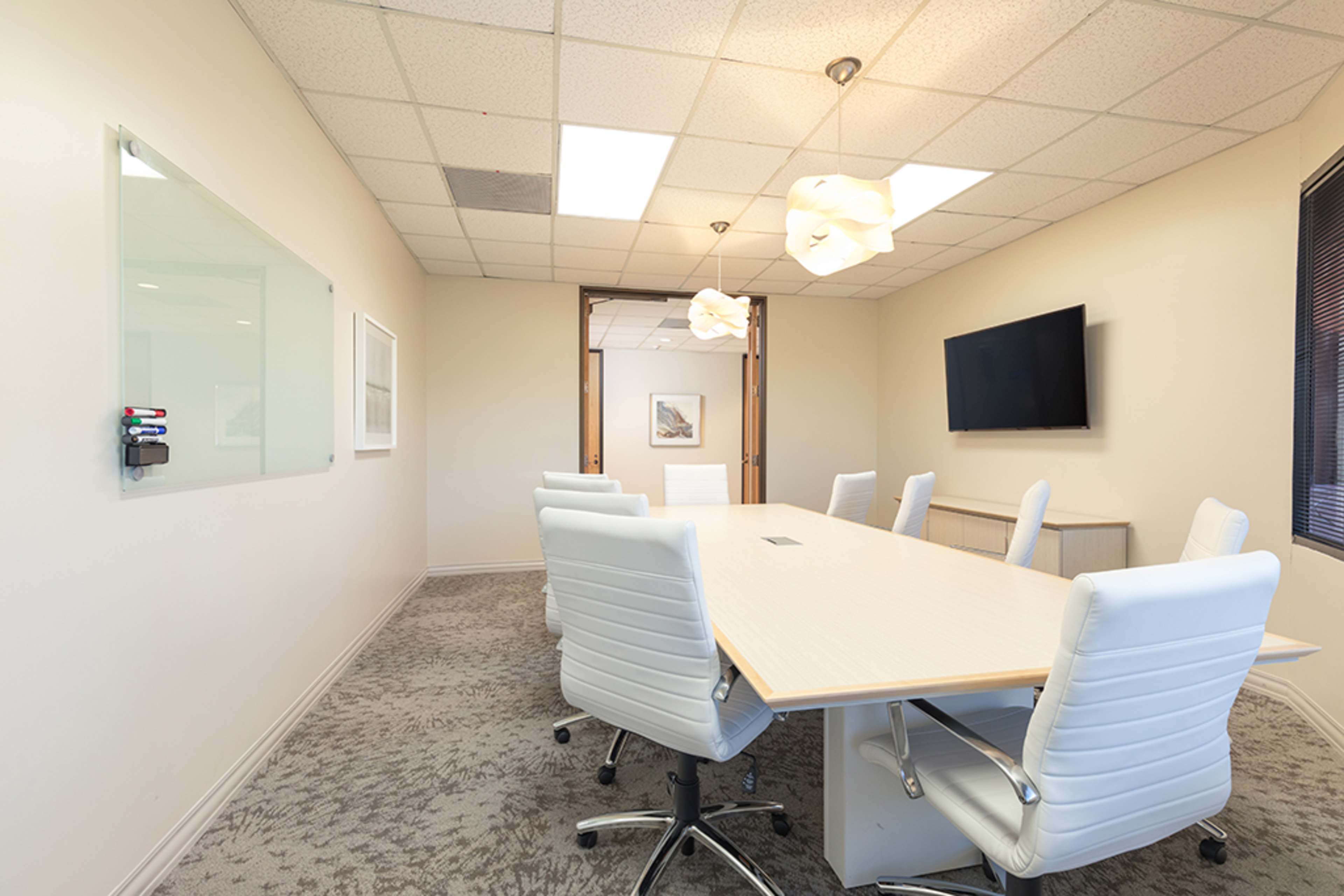 A modern conference room features a long table surrounded by white ergonomic chairs, with a wall-mounted TV and overhead lighting.