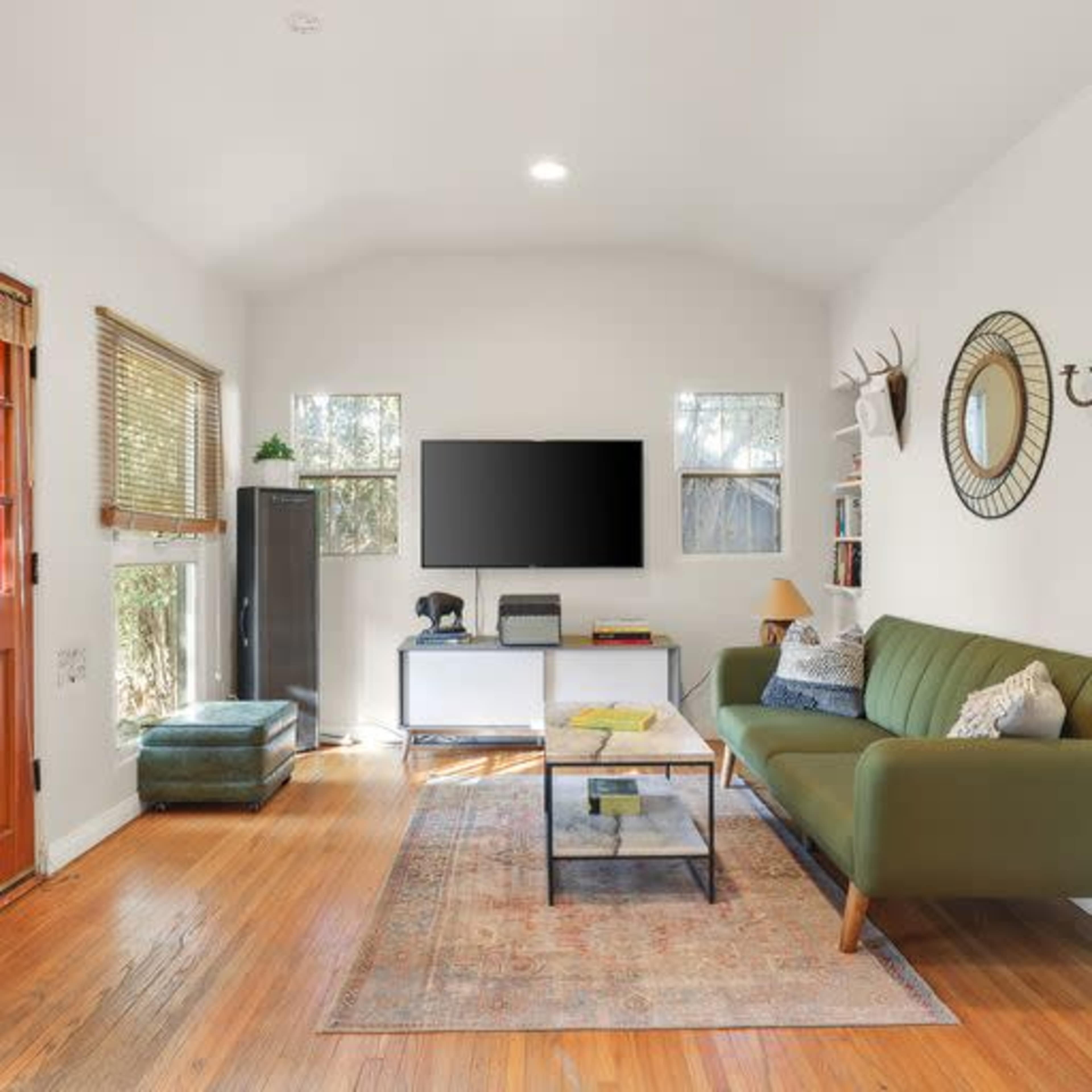 A small living room with a green sofa, a coffee table, a television on a white stand, and a few windows allowing natural light.