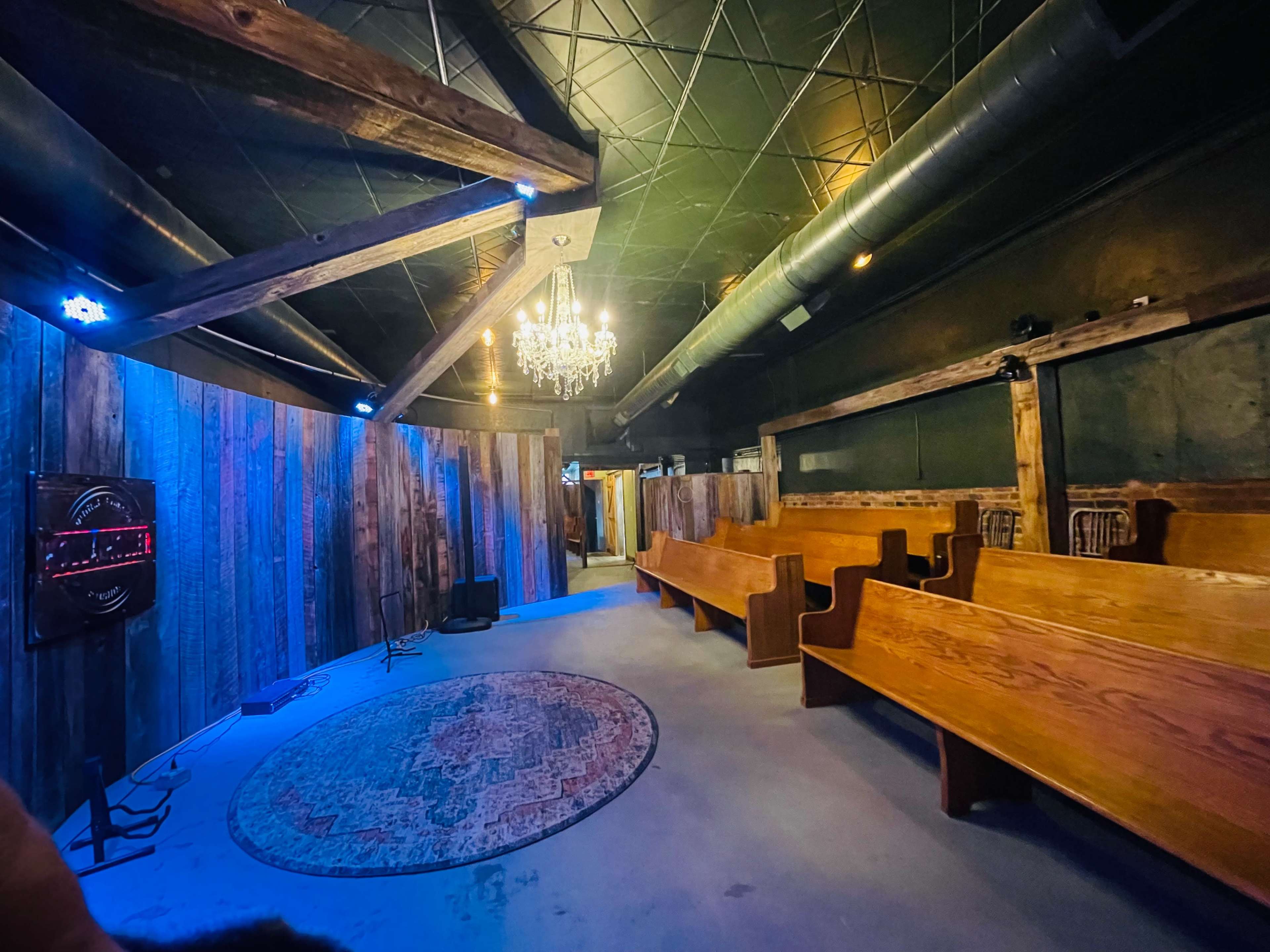 The image shows a dimly lit interior space with wooden benches, a circular rug, and a chandelier hanging from the ceiling.