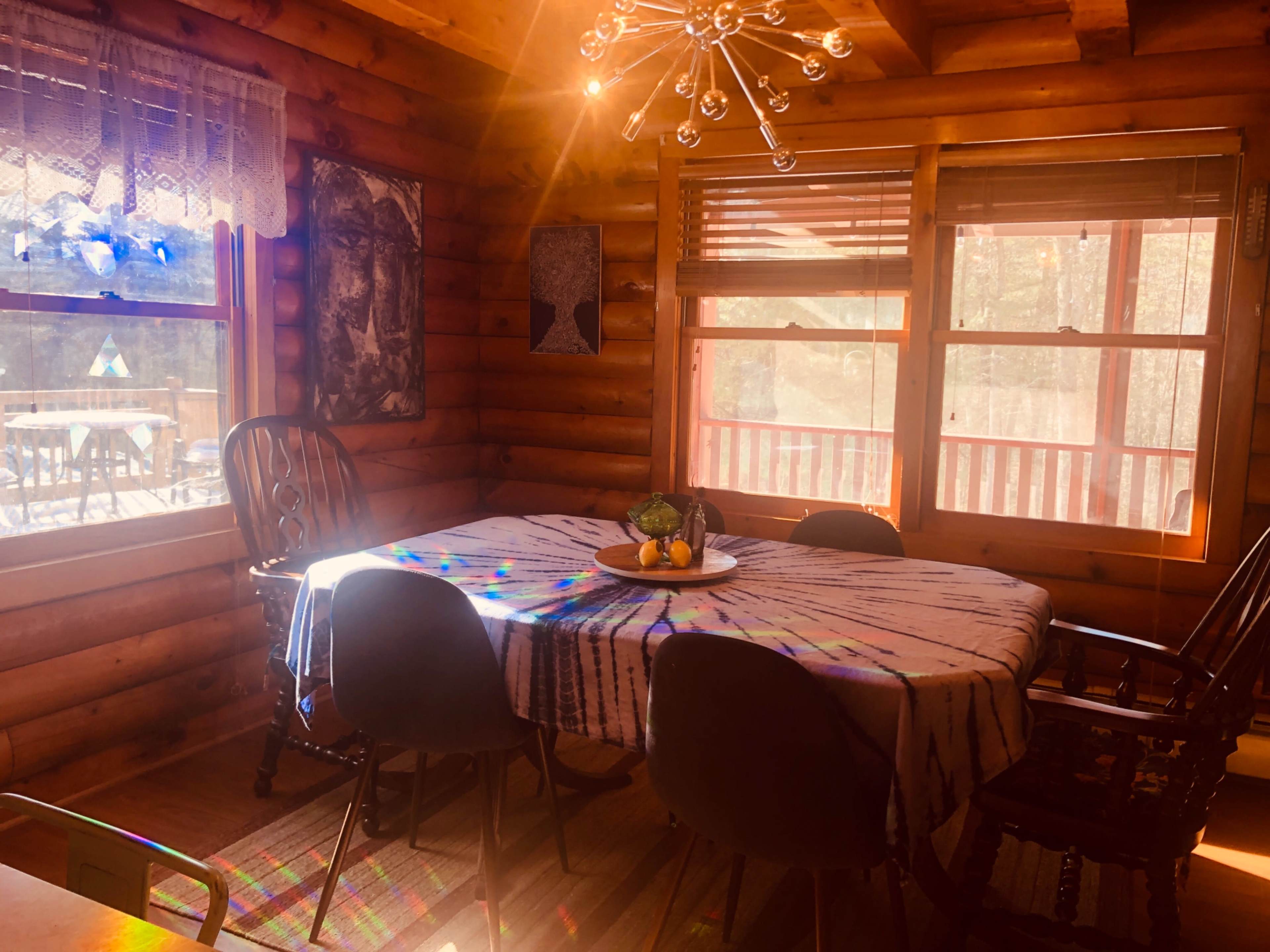 A wooden dining area features a table covered with a tie-dye tablecloth, surrounded by chairs and illuminated by a modern chandelier.