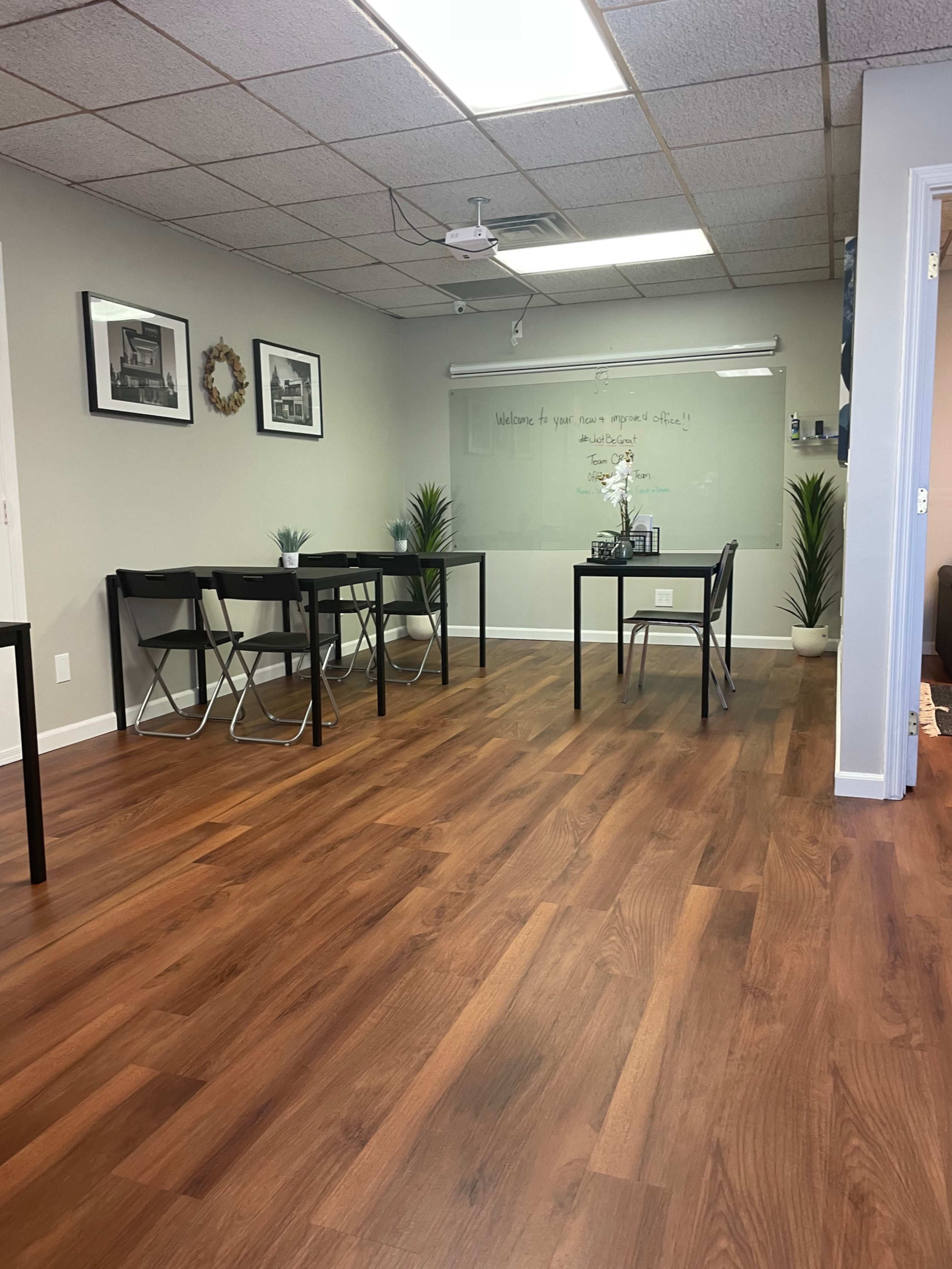A well-lit room with wooden flooring, featuring black folding tables and chairs, and plants in the corners.