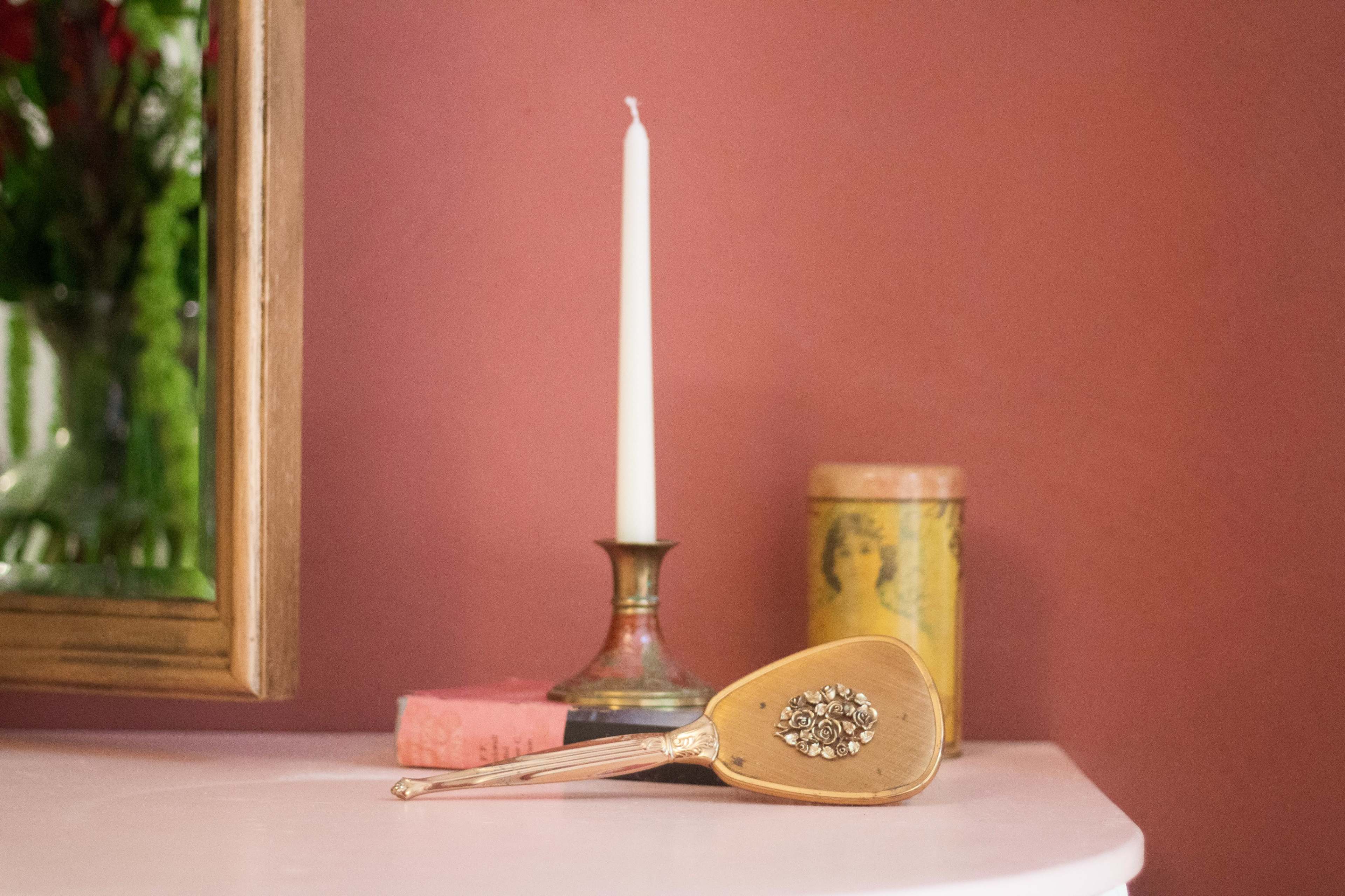 A vintage hand mirror with a floral design rests on a white table near a candle and decorative containers against a pink wall.