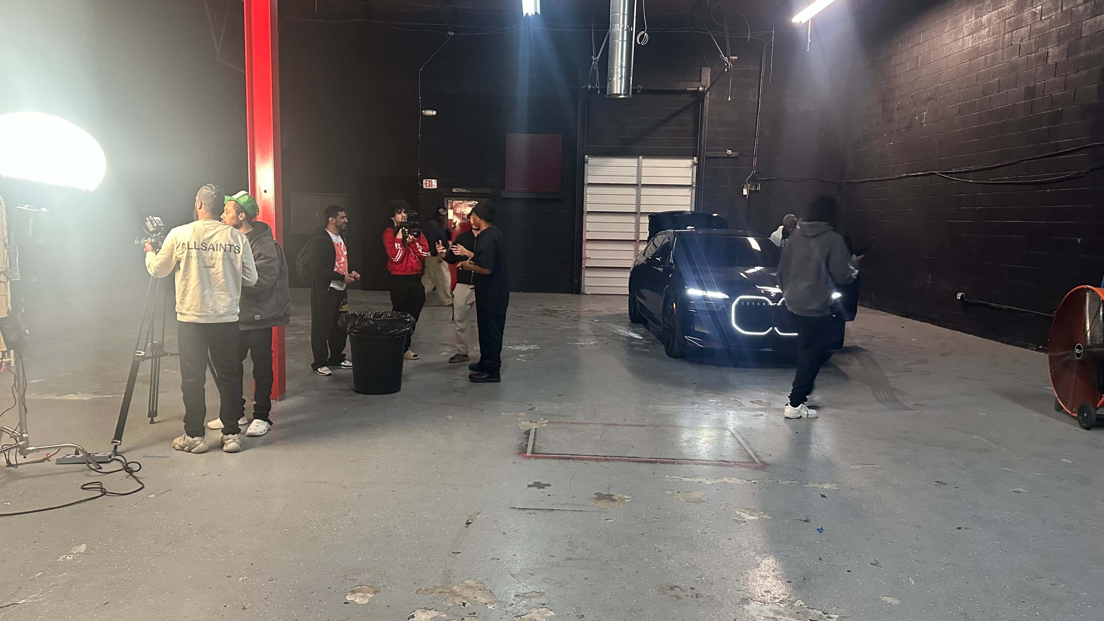 A group of people is gathered in a dimly lit warehouse, with a dark car illuminated by lights in the foreground.