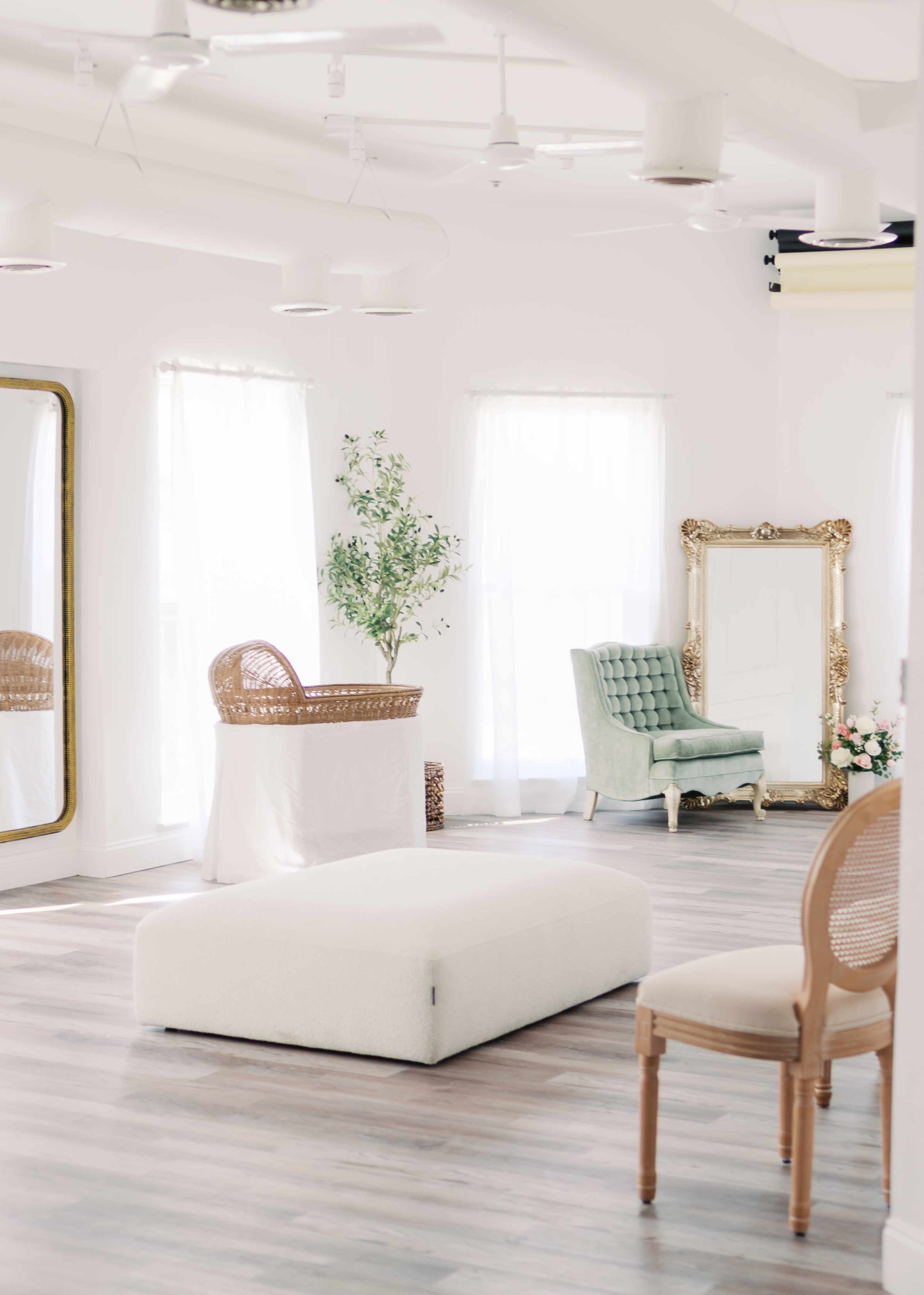 The image shows a bright, airy room with large windows, featuring a green upholstered chair, a large mirror, and minimalist furniture.