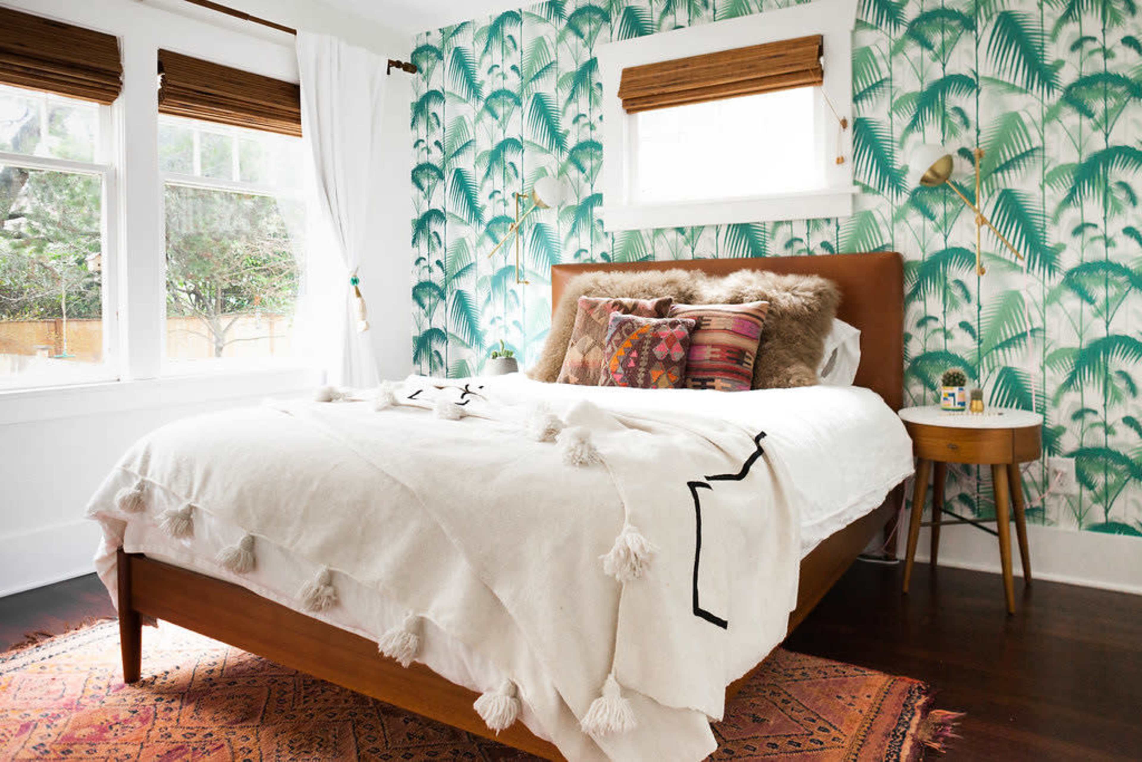 This bedroom features a bed with a light blanket and decorative pillows, surrounded by palm-patterned wallpaper and a nightstand with a lamp.