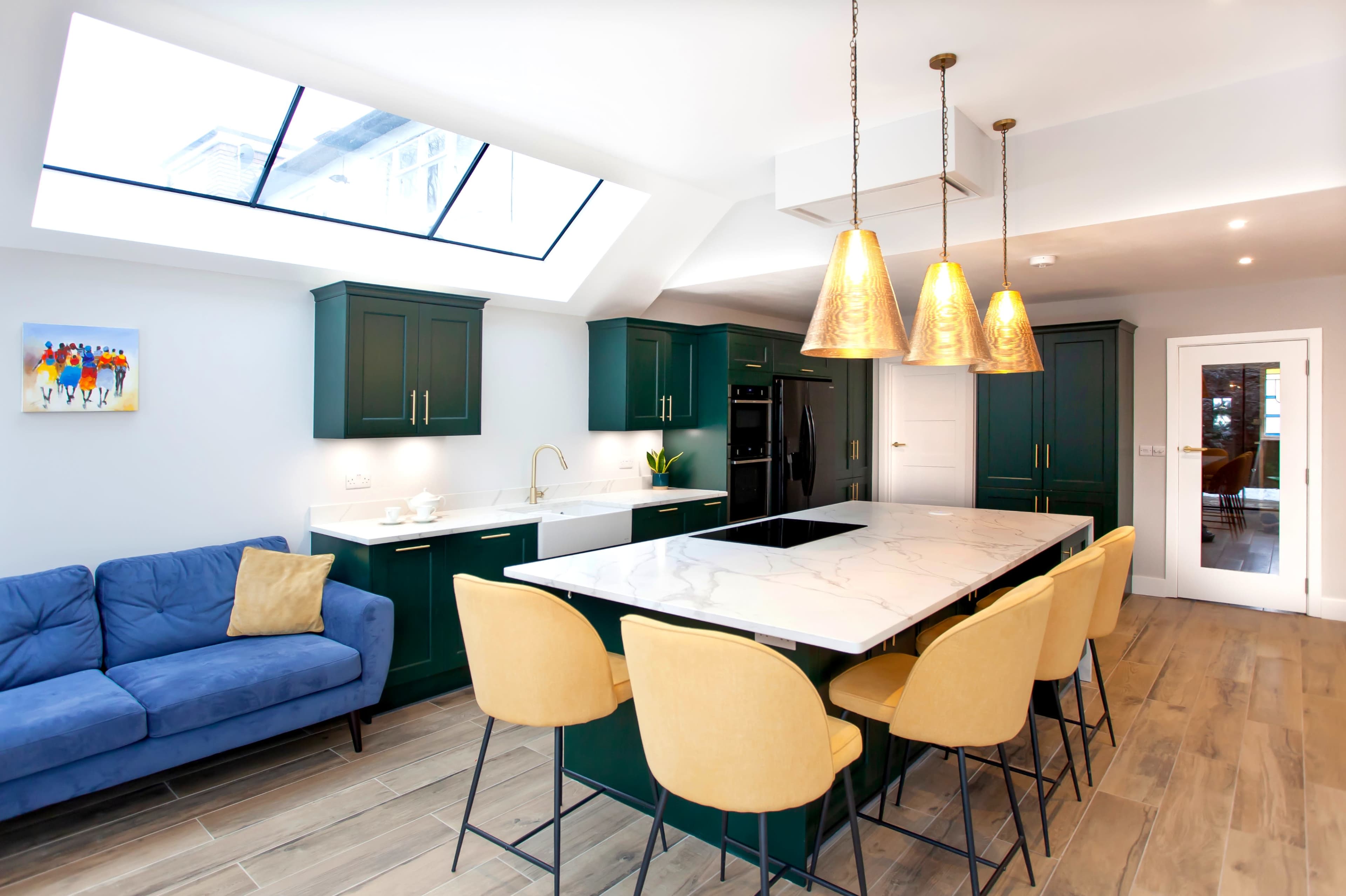 The kitchen features green cabinetry, a large marble island with yellow chairs, and a skylight above.