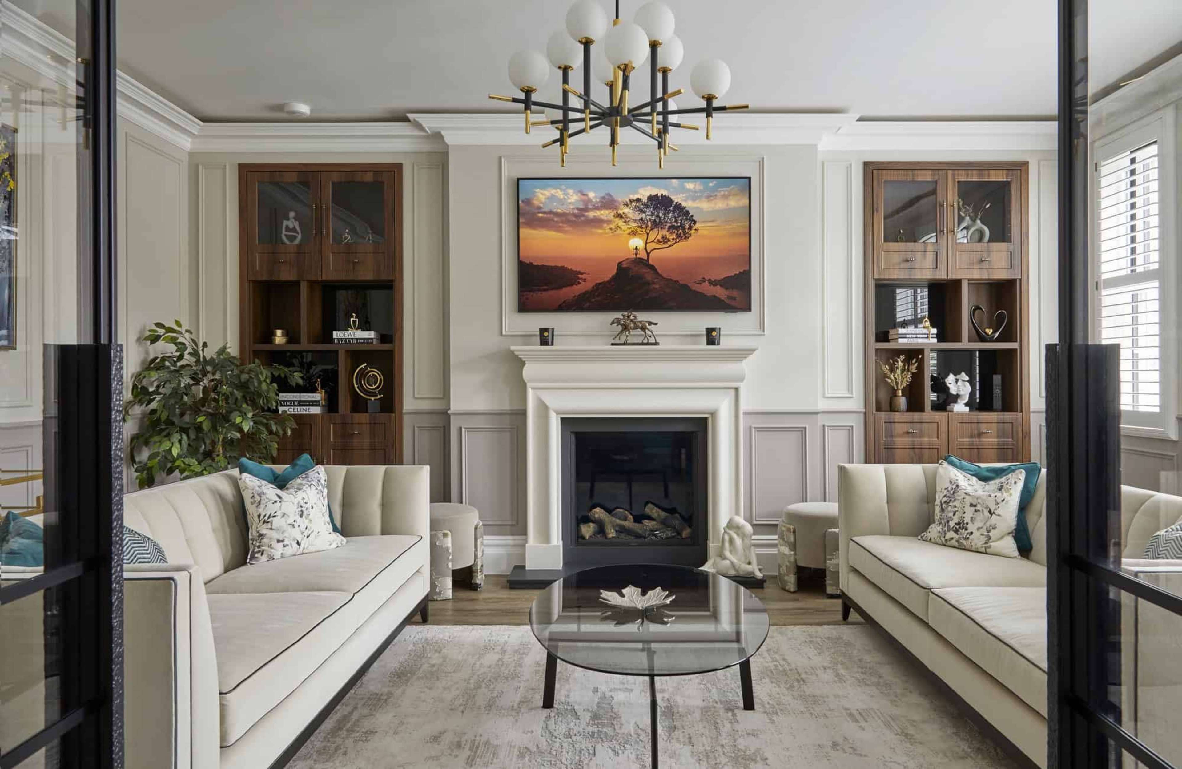 A modern living room features two white sofas, a glass coffee table, a television mounted above a fireplace, and wooden bookcases on either side, all framed by decorative wall paneling.
