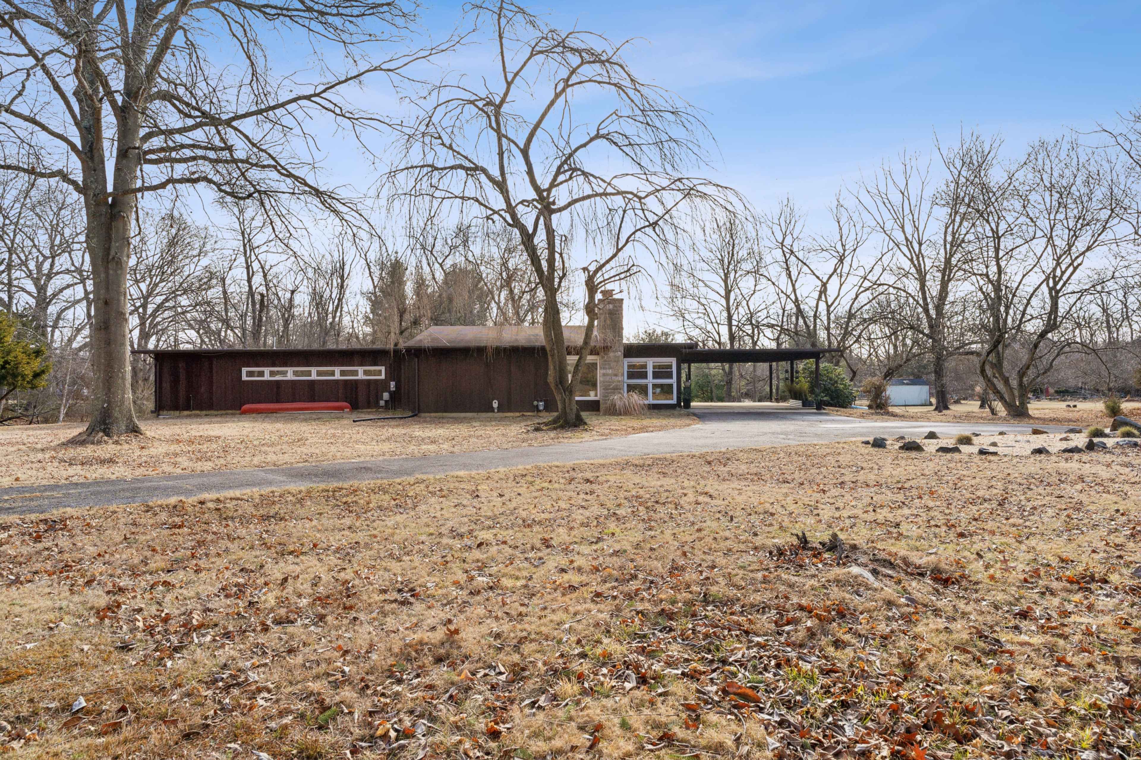 The image shows a modern-style house with large windows, situated on a grassy lot surrounded by bare trees in early spring.