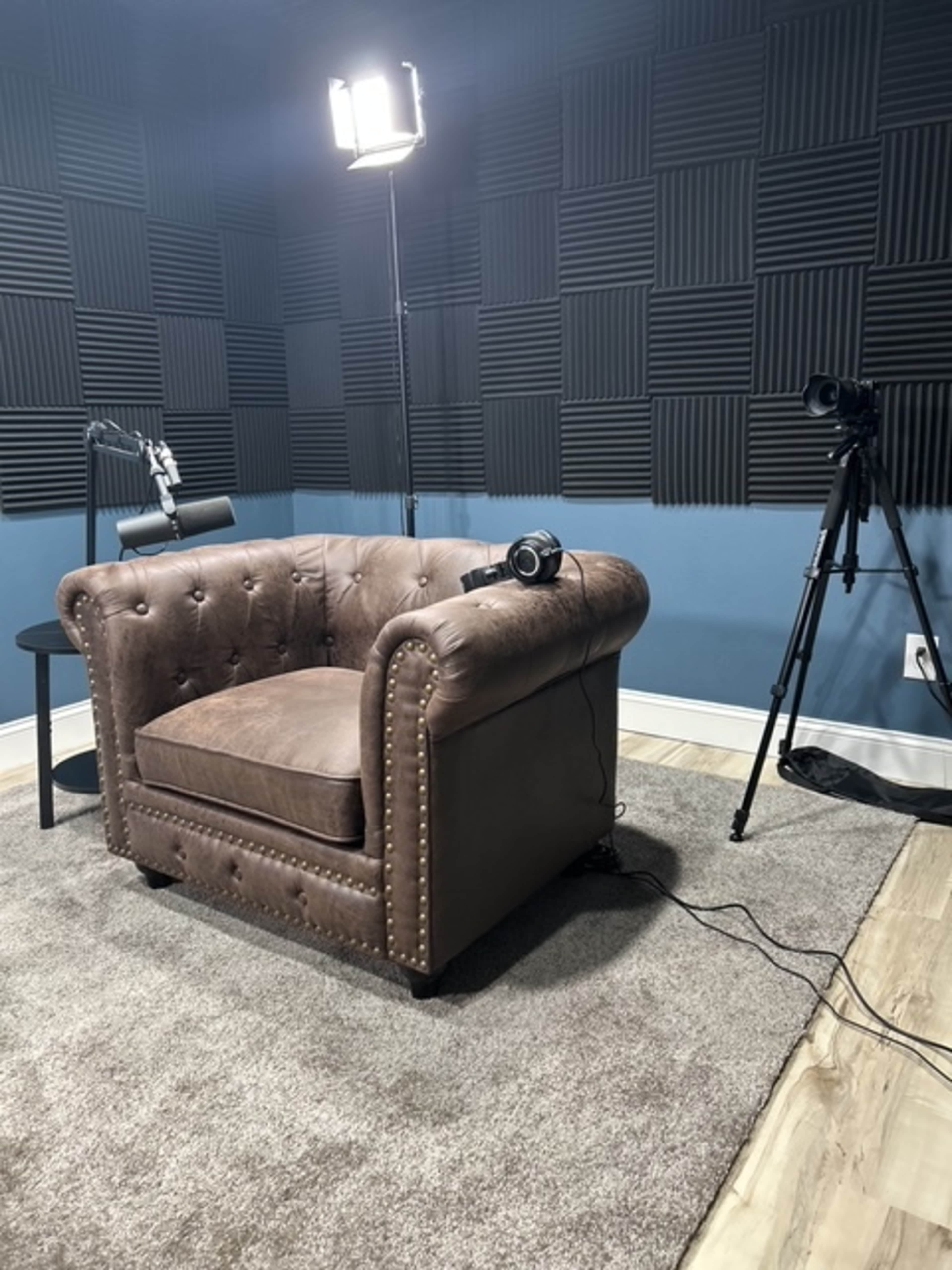 The image shows a brown leather armchair positioned in a room with acoustic wall panels, a spotlight overhead, and a tripod camera nearby.