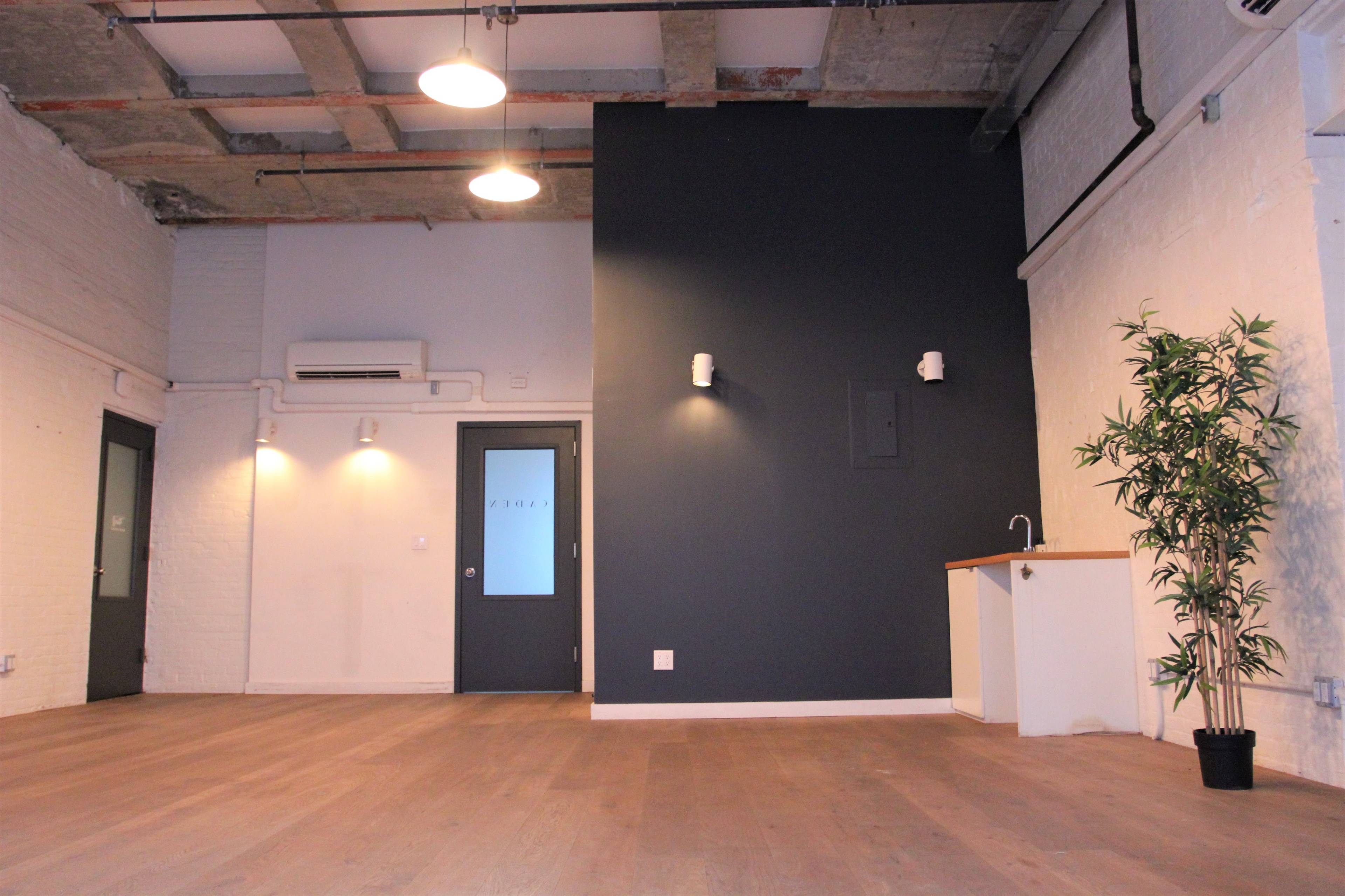 An empty room with wooden flooring, a dark wall, a small kitchenette area, and a door at the far end.