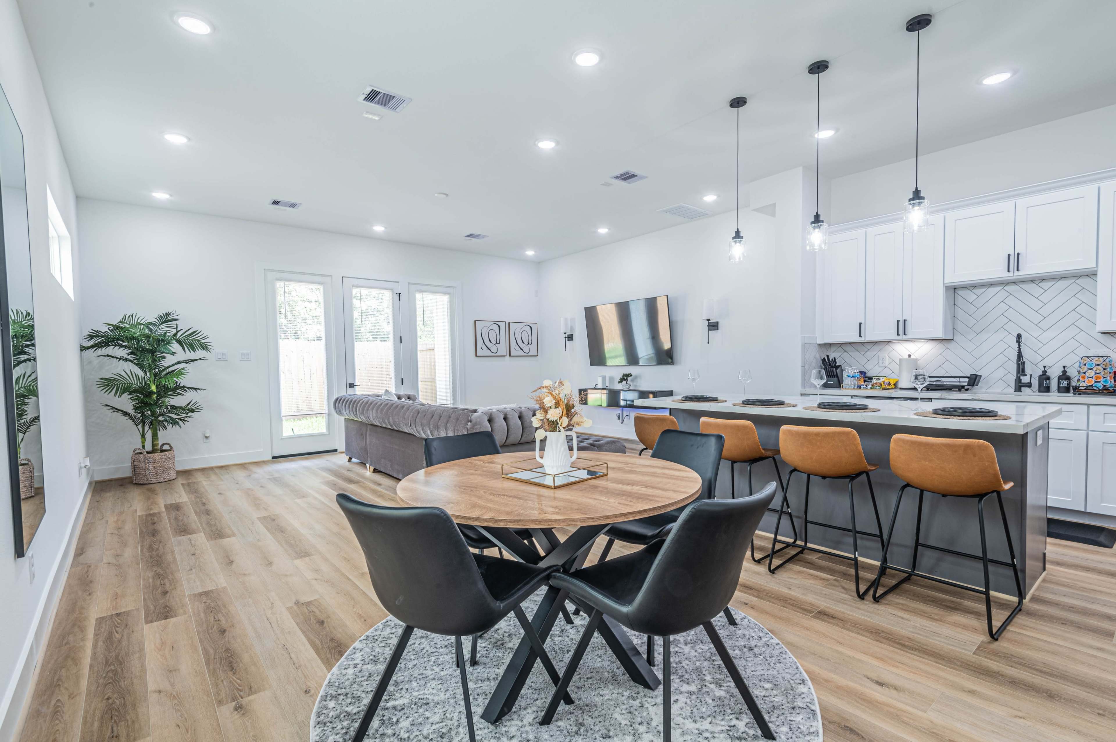 A modern living space featuring a dining area with a round table and black chairs, a kitchen with white cabinetry, and a cozy seating area with a large couch and a television.