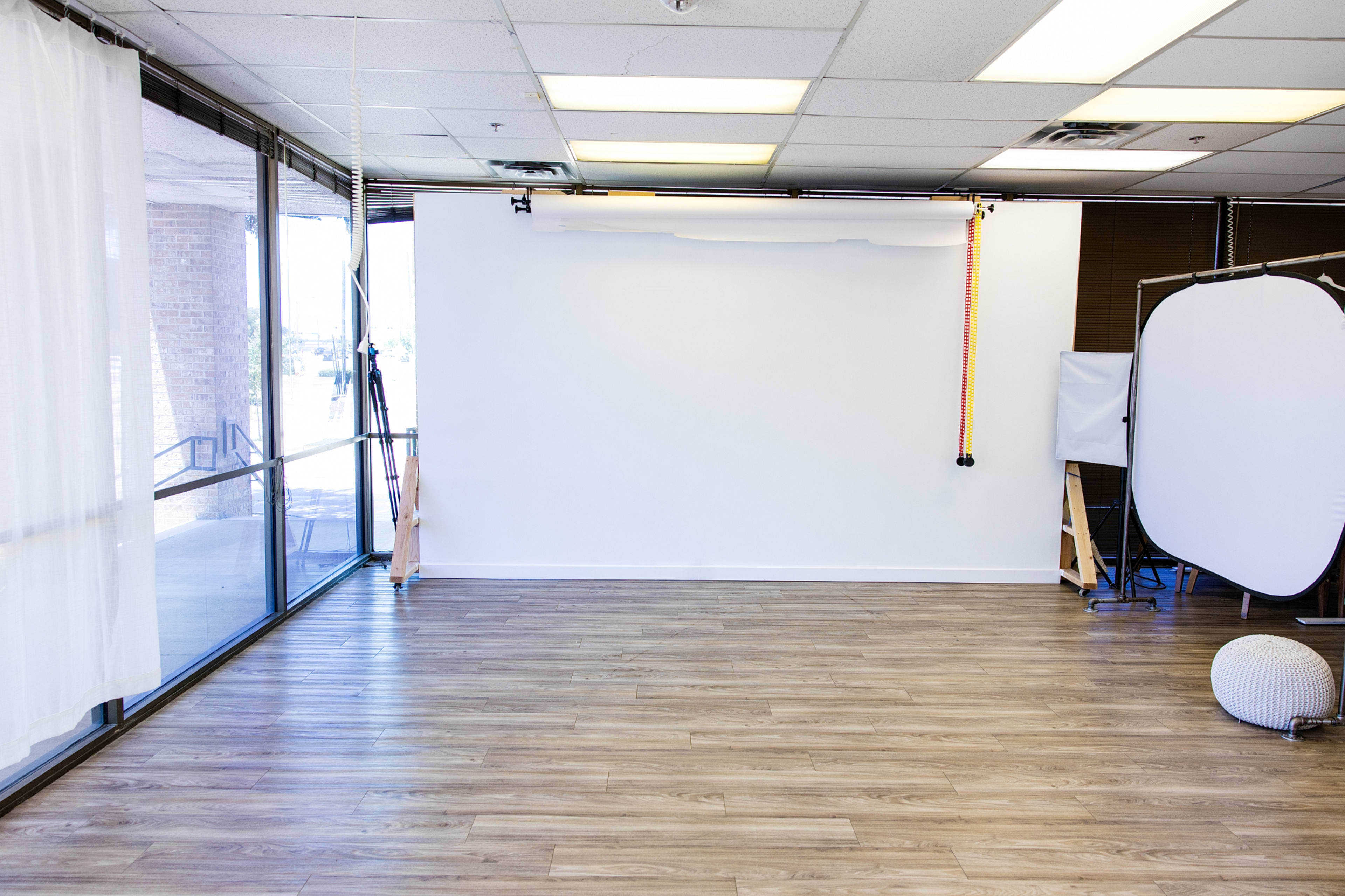 The image shows a spacious, empty room with large windows and a white backdrop setup for photography.