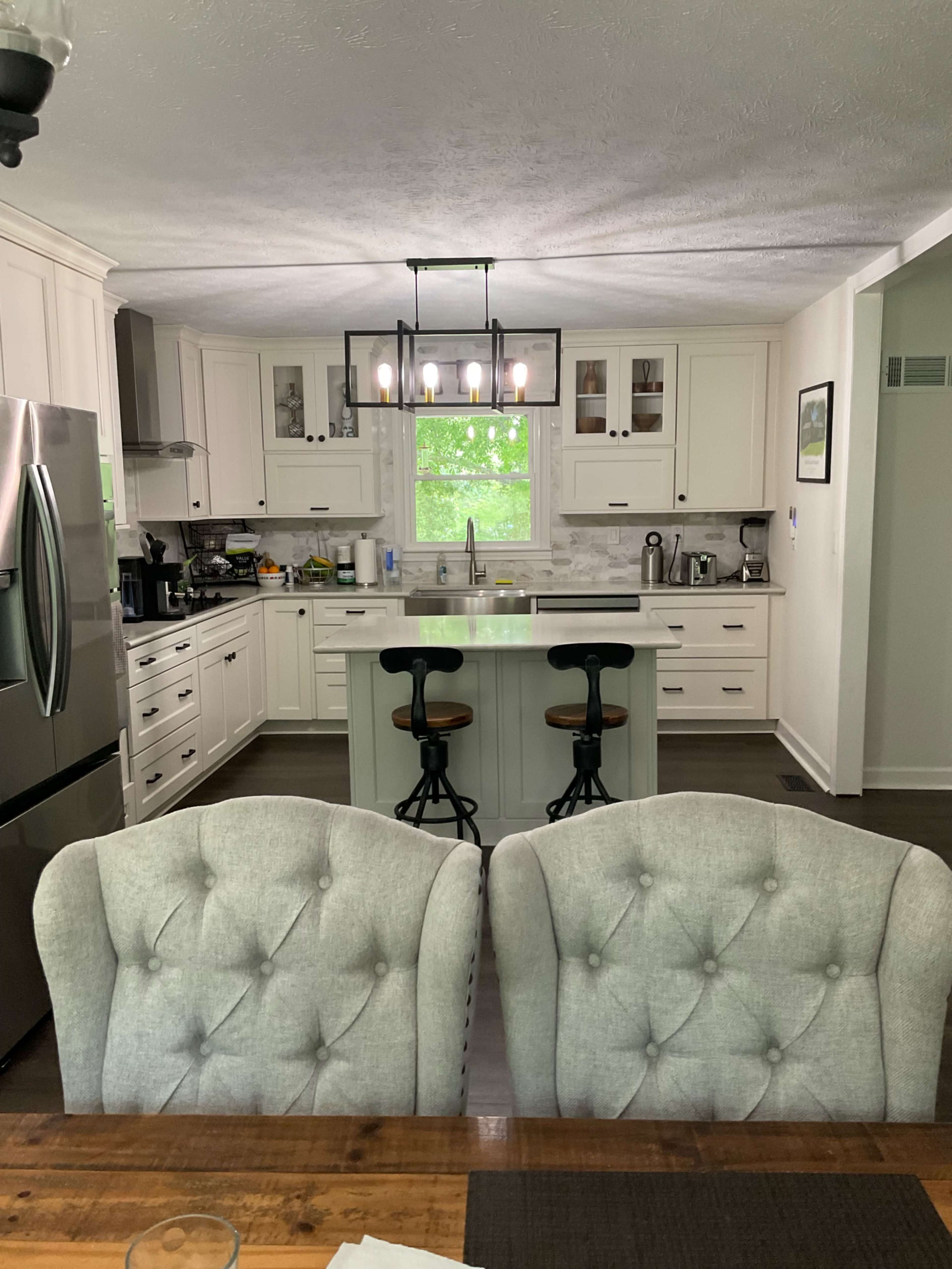 A modern kitchen features white cabinets, a central island with bar stools, and a view of a green outdoor area through a window.