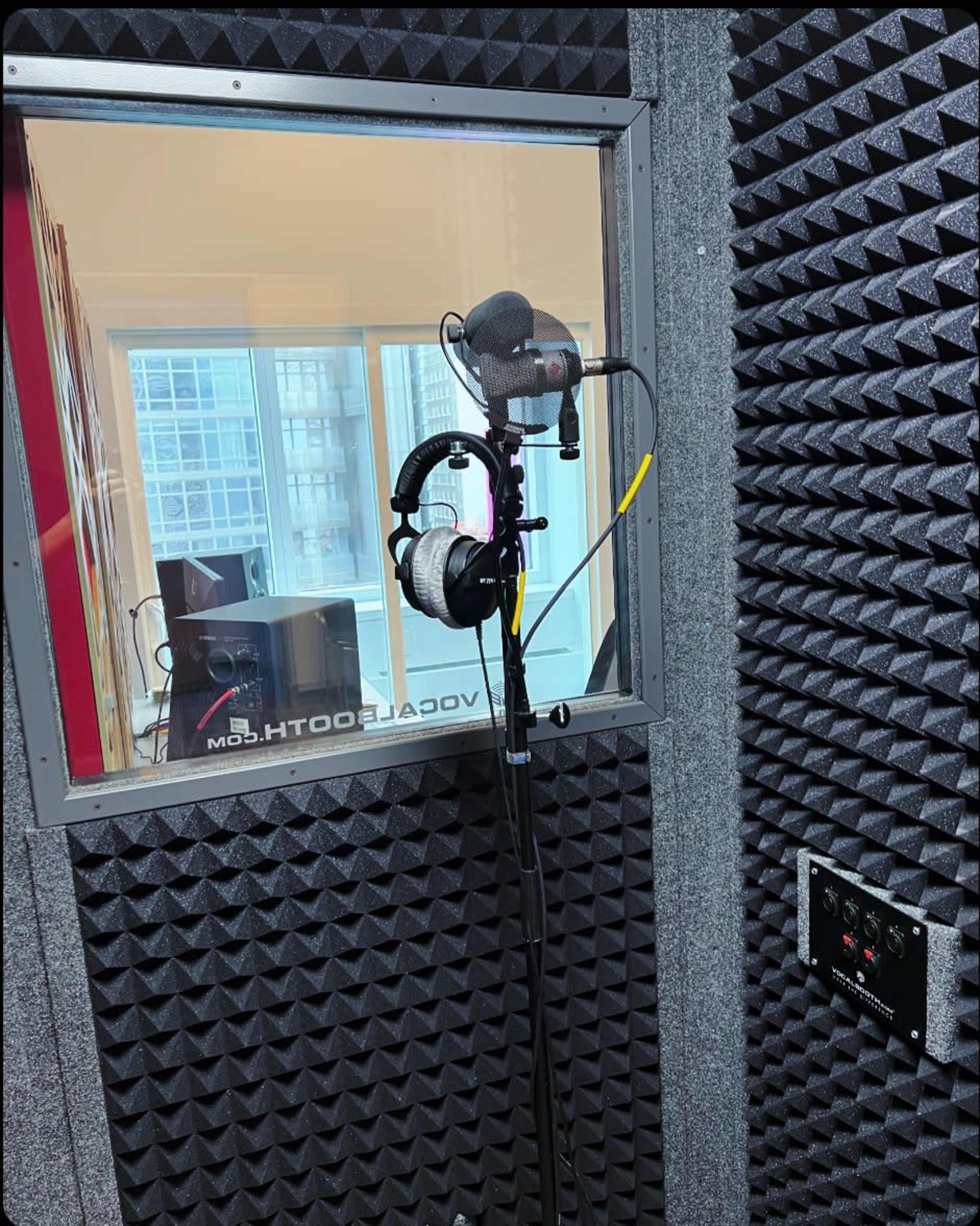 A microphone stands on a stand with headphones hanging from it inside a soundproof recording studio booth.