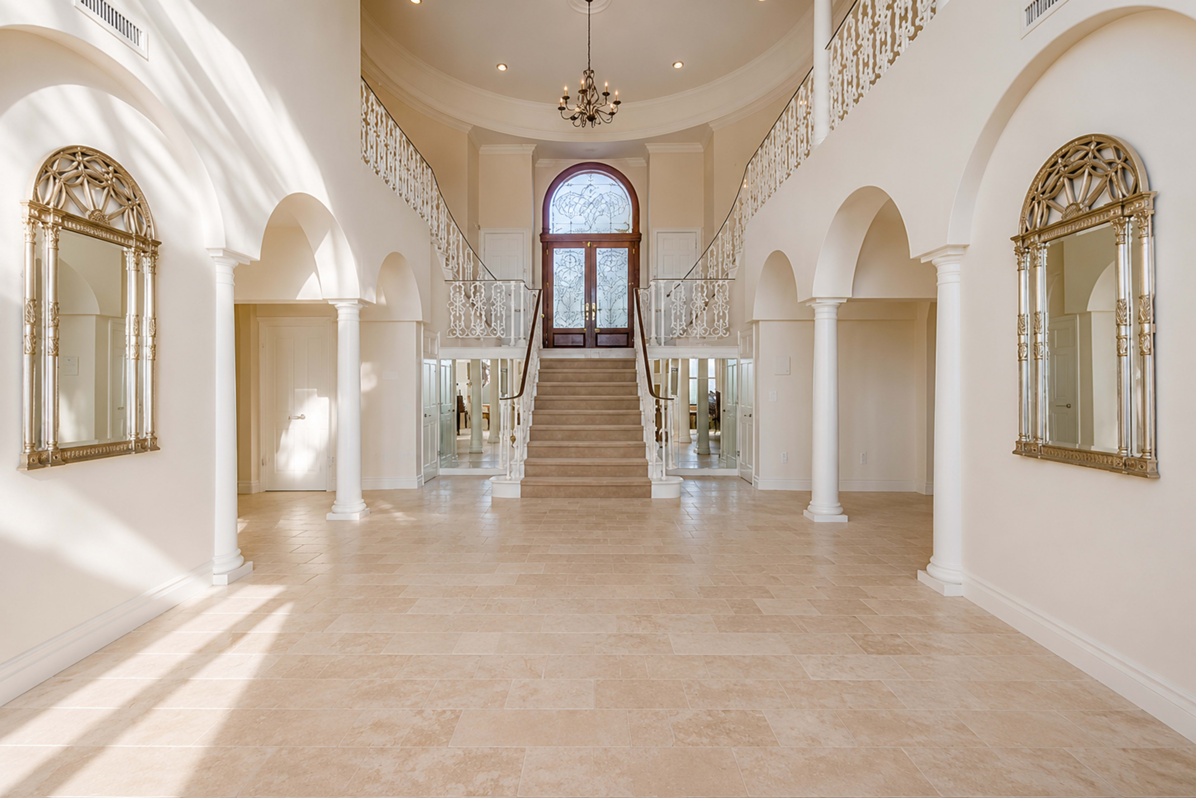 The image shows a large, airy foyer with a grand staircase, ornate mirrors, and a chandelier, illuminated by natural light streaming through large windows.