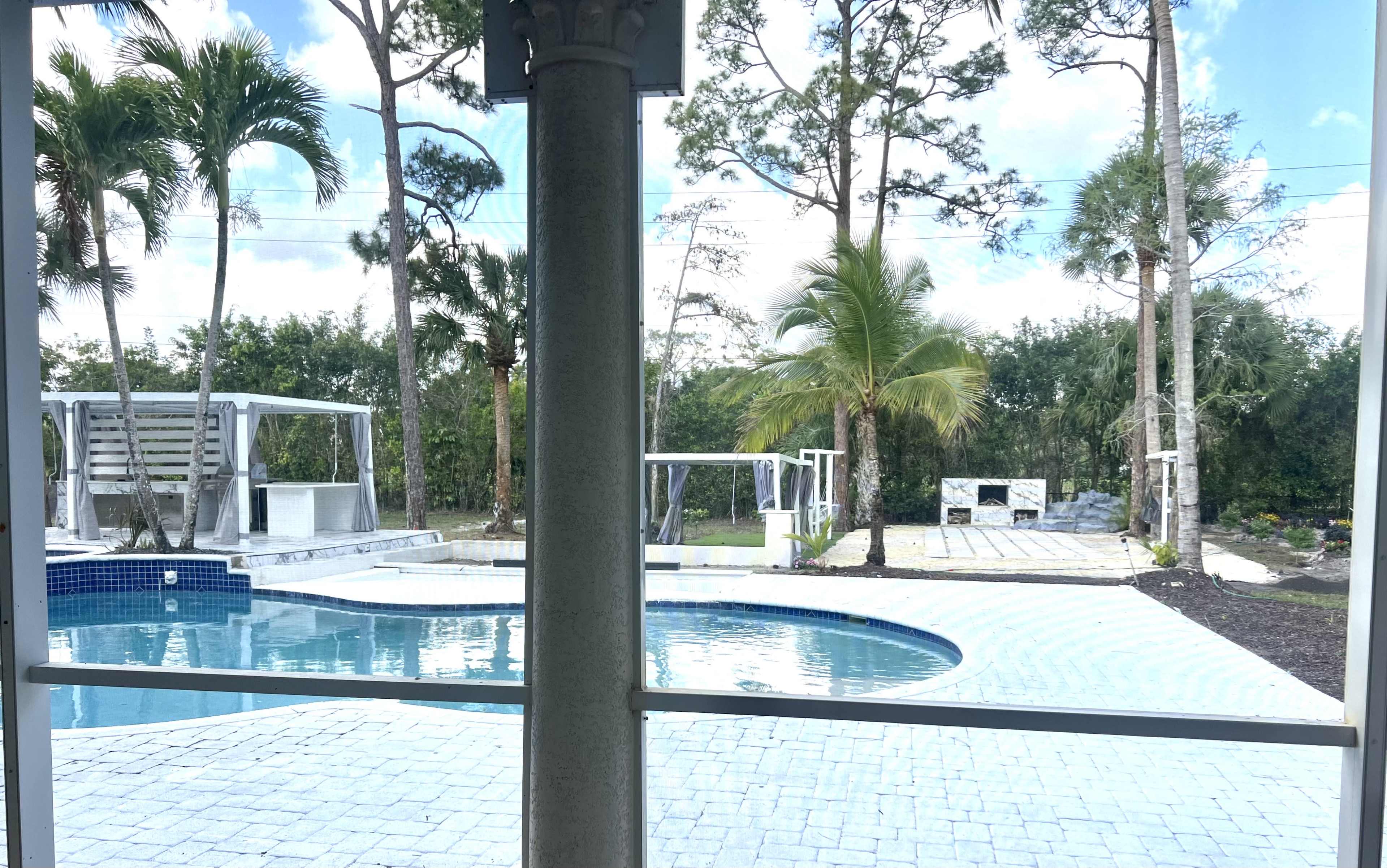 The image shows a backyard with a swimming pool, surrounded by palm trees and outdoor structures, including seating areas and a built-in barbecue workstation.