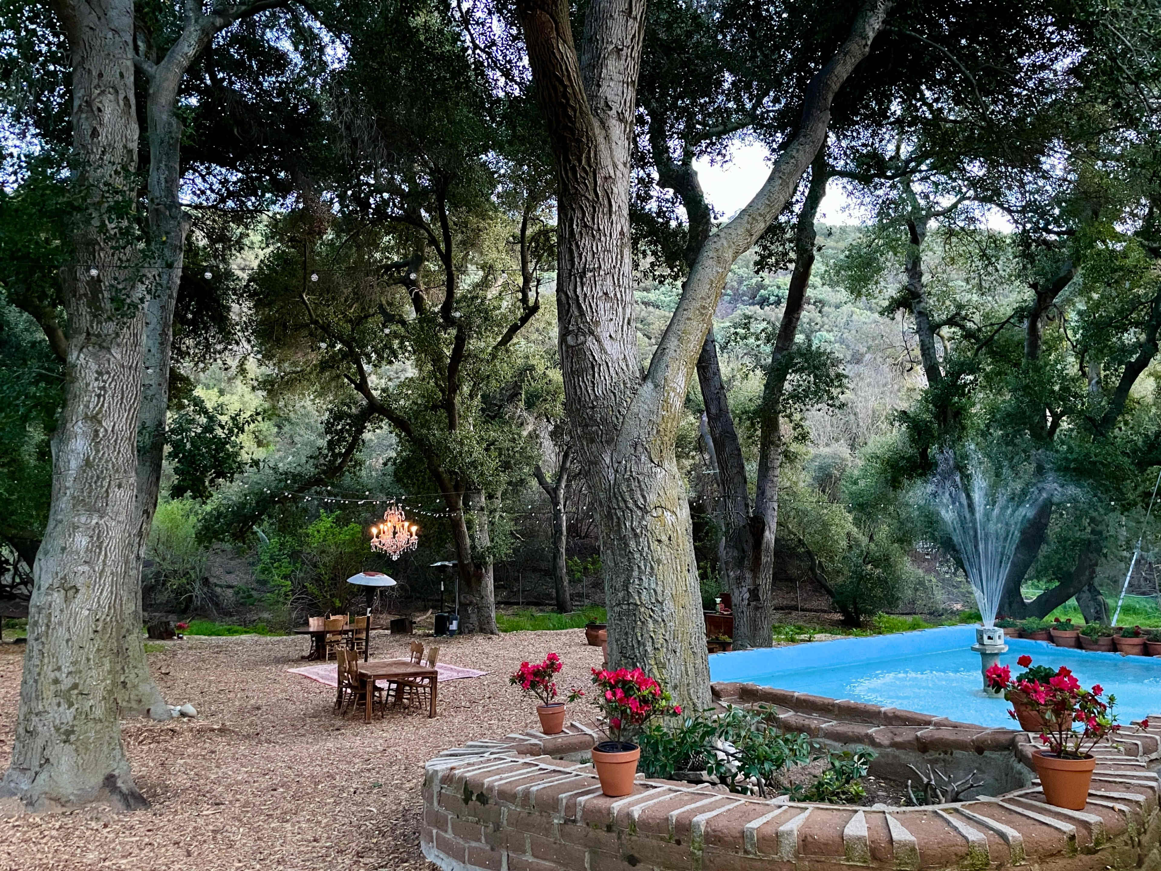 A landscaped outdoor area features a blue fountain surrounded by potted flowers, with dining tables set beneath trees and a chandelier hanging nearby.