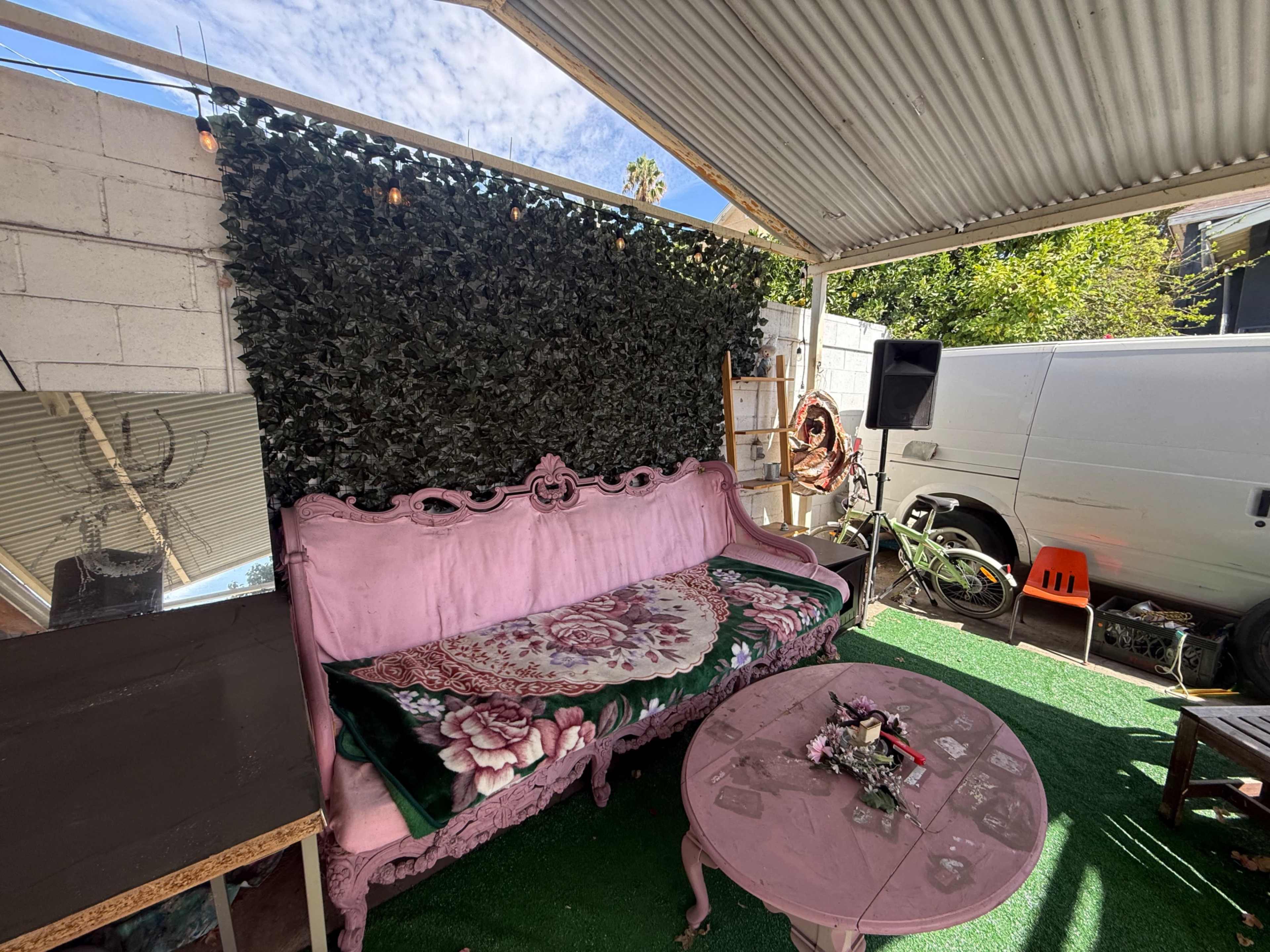 The image shows a cozy outdoor seating area with a pink vintage couch, a round table, and a green artificial grass floor beneath a metal roof.