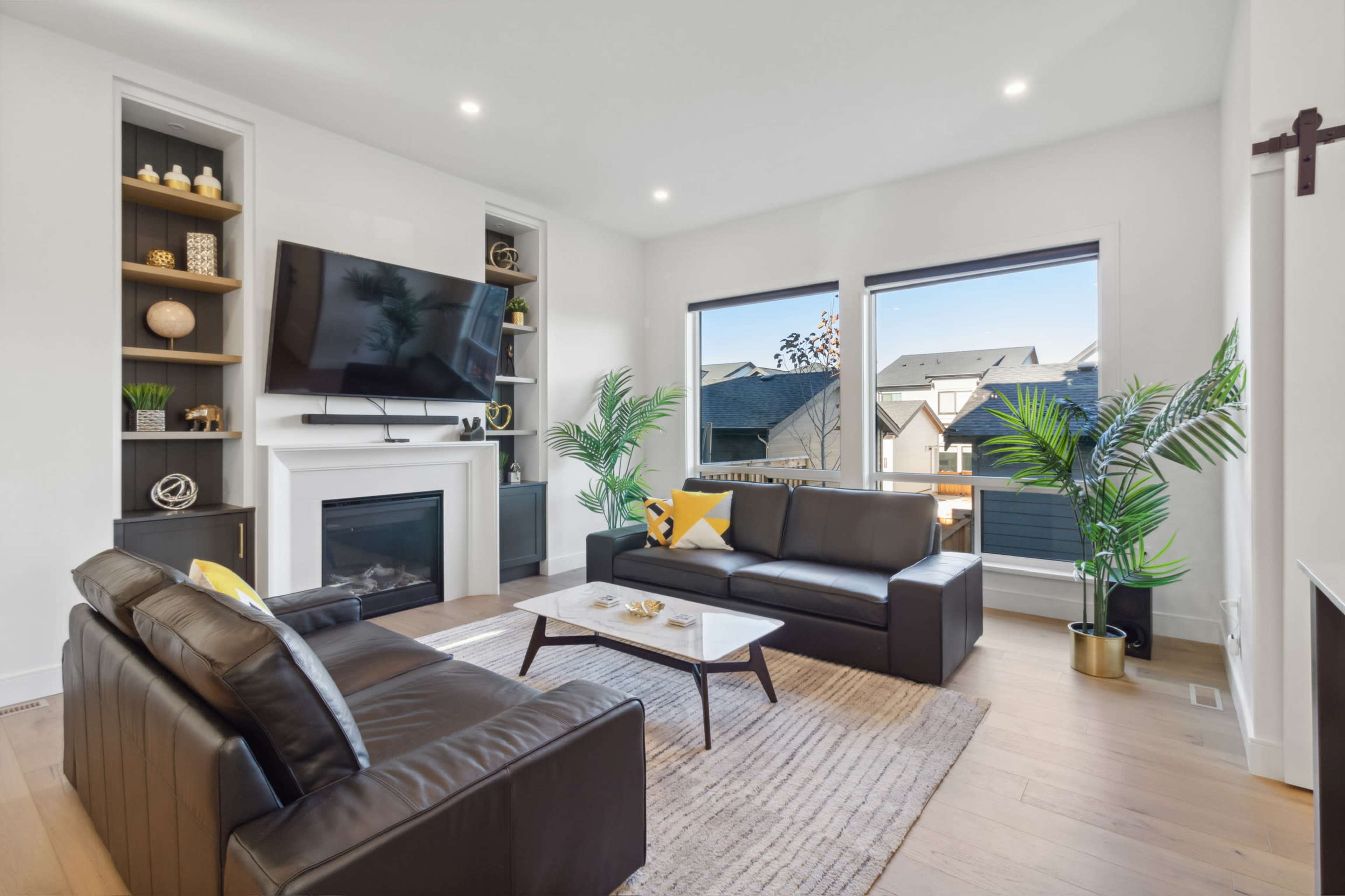 A modern living room features a fireplace, large windows with outdoor views, and contemporary furniture arranged around a coffee table.