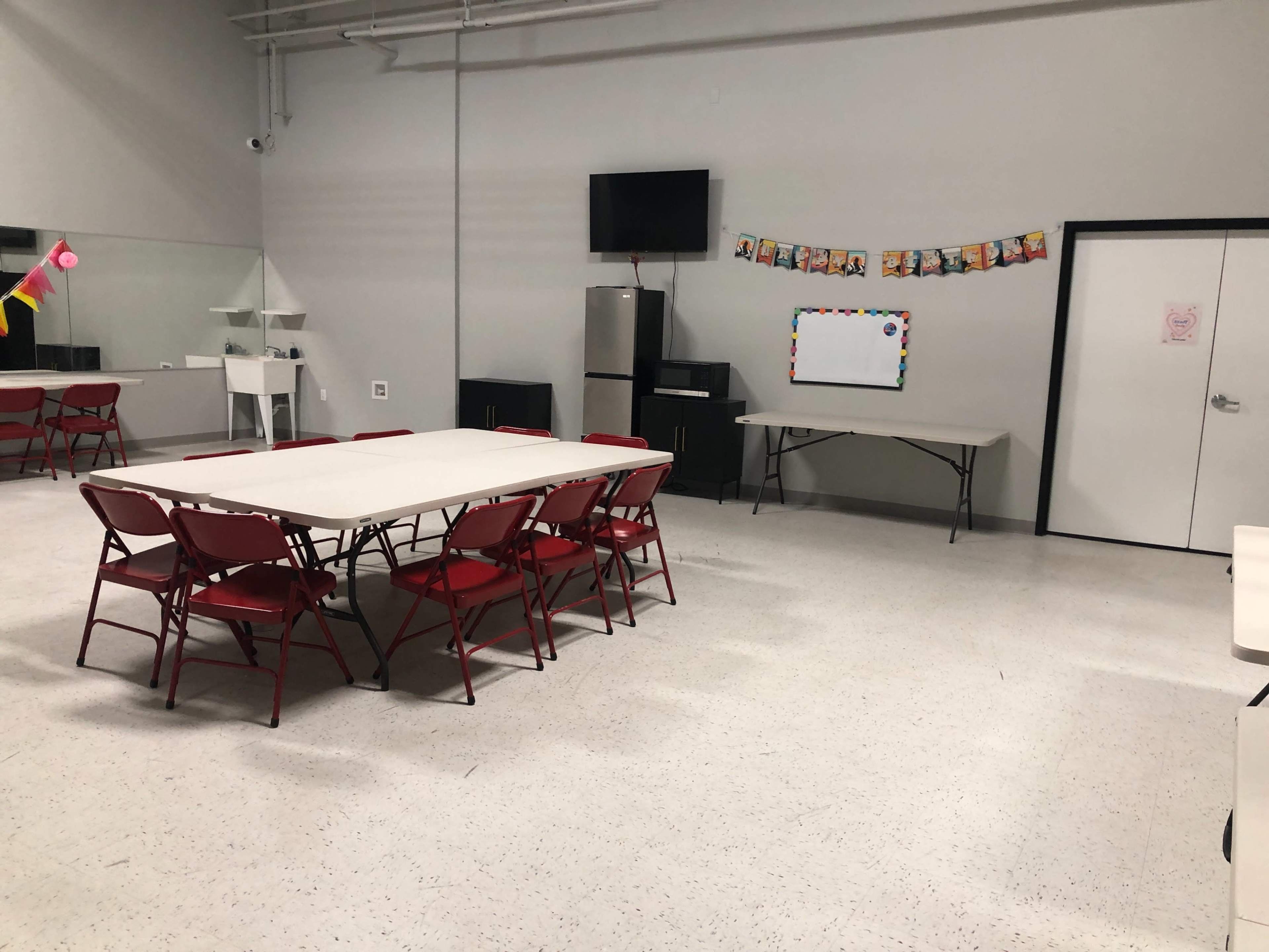 The image shows a spacious room with a large table surrounded by red folding chairs, a television mounted on the wall, a refrigerator, and a blank whiteboard next to a decorated wall.