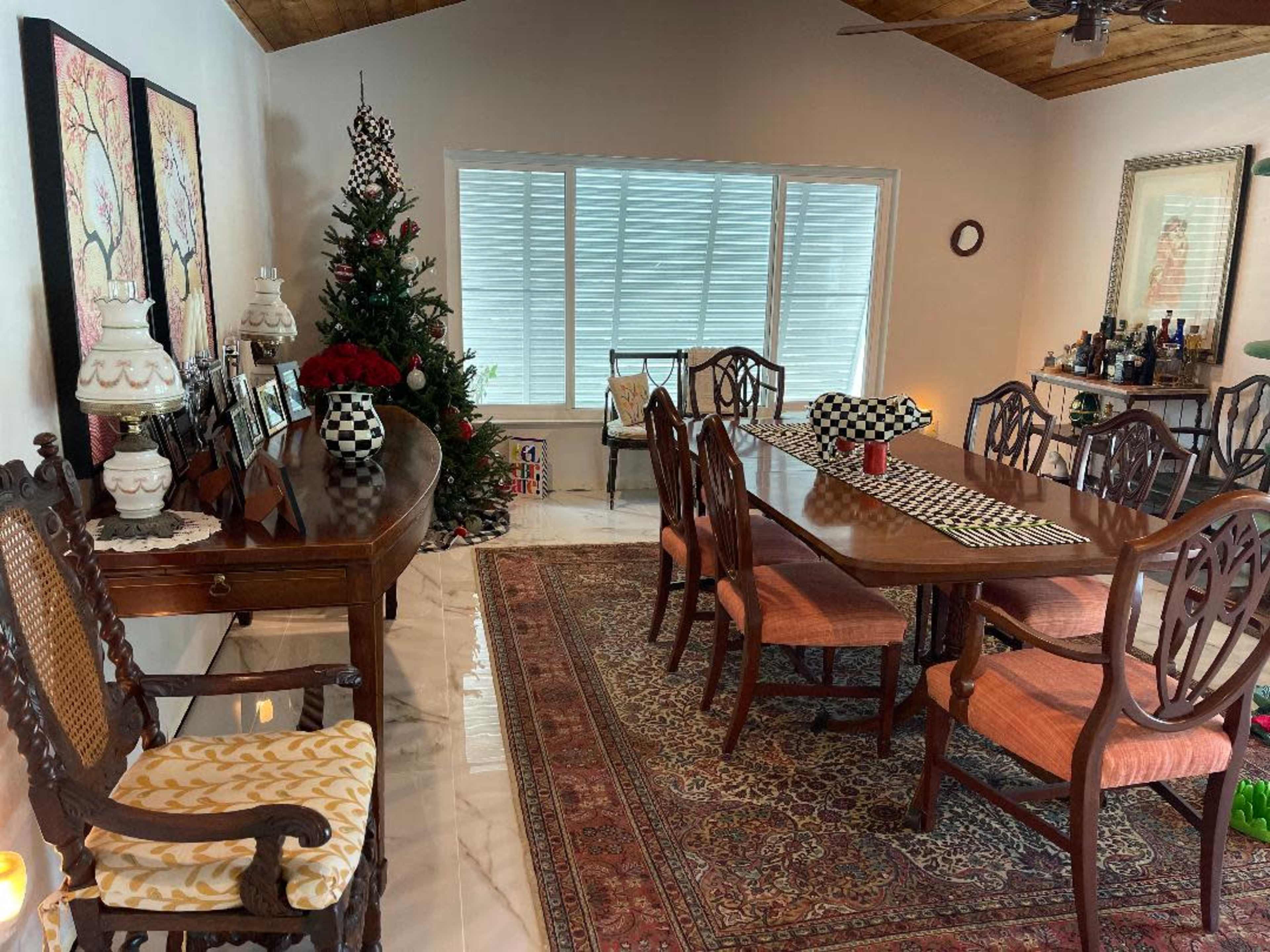 The image shows a dining area with a wooden table, several chairs, a Christmas tree in the corner, and large windows allowing natural light to enter.