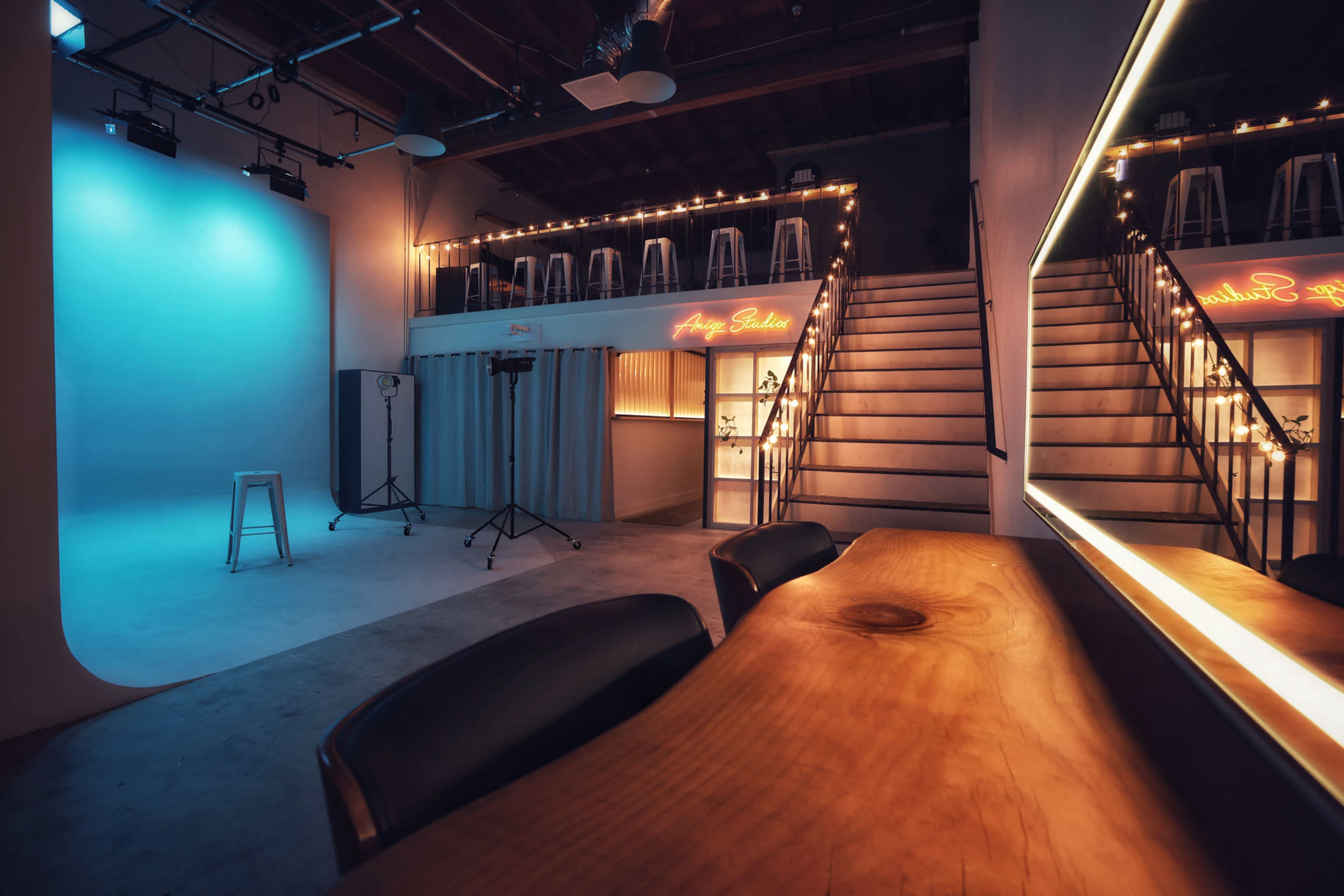 A studio space featuring a bright blue backdrop, a wooden table with black chairs, and a staircase leading to an upper level decorated with string lights.