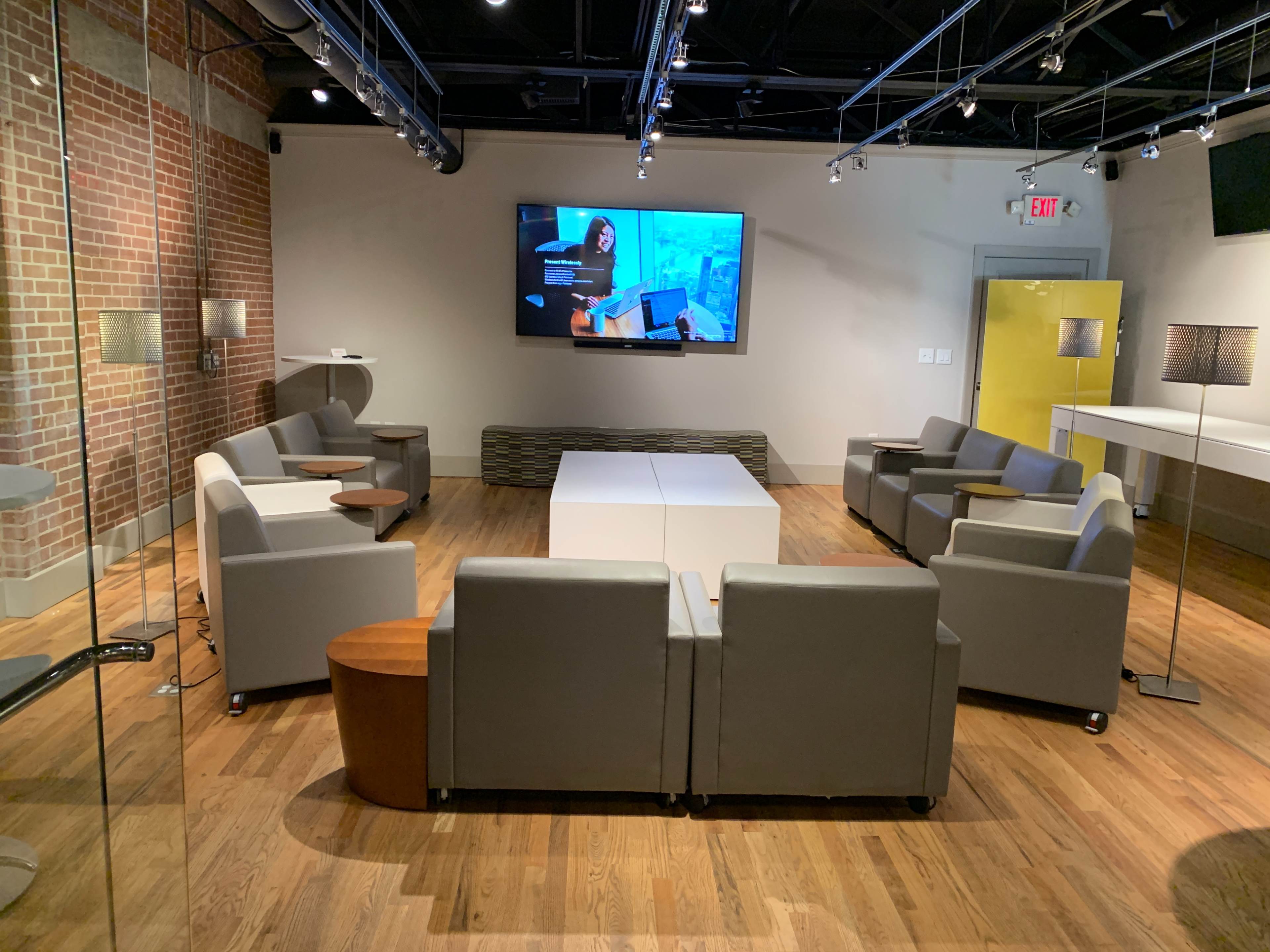 A modern lounge area with gray seating, a large screen on the wall, and wooden flooring.
