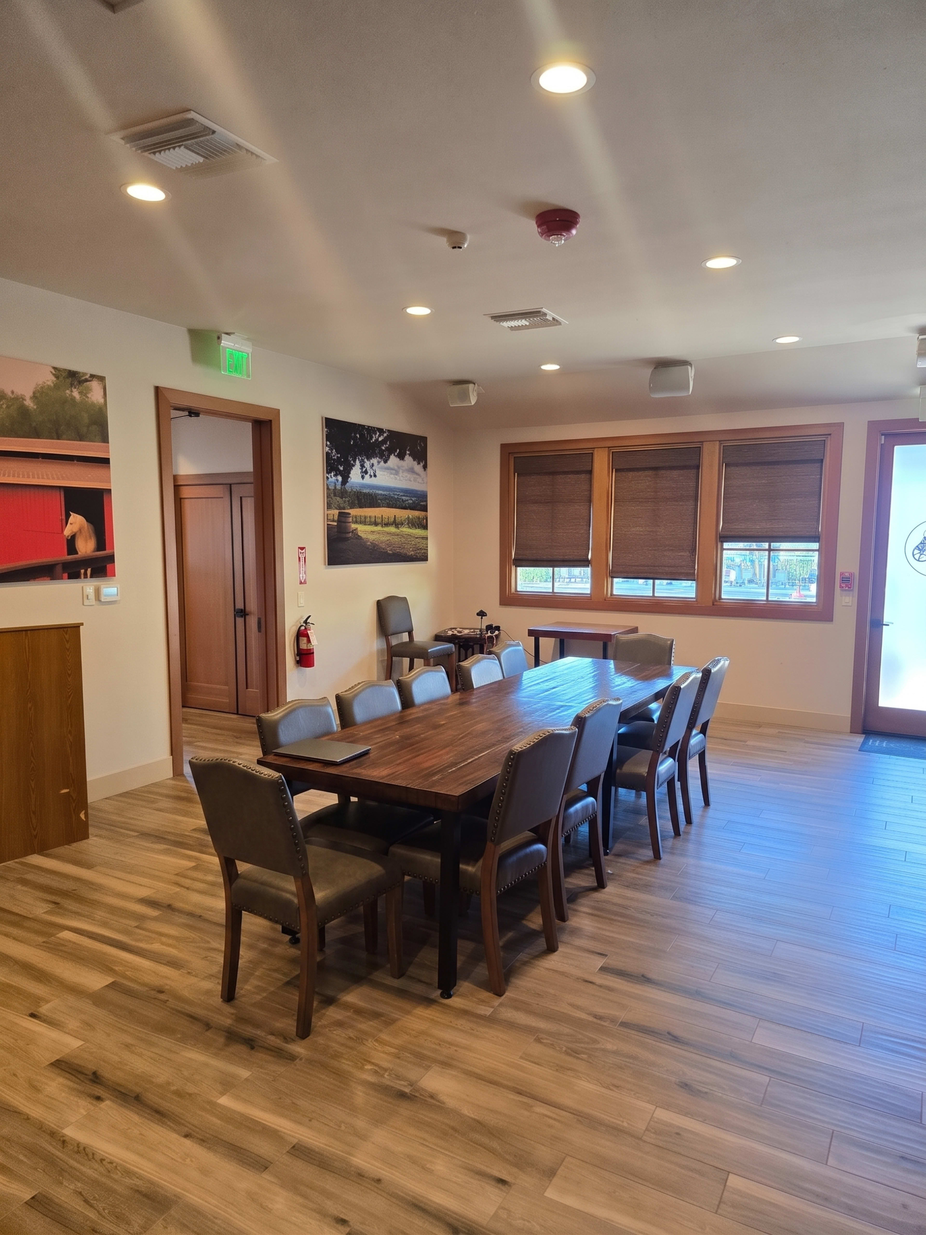 The image shows a well-lit meeting room with a long wooden table surrounded by chairs, along with a side wall featuring a large painting and windows with shades.