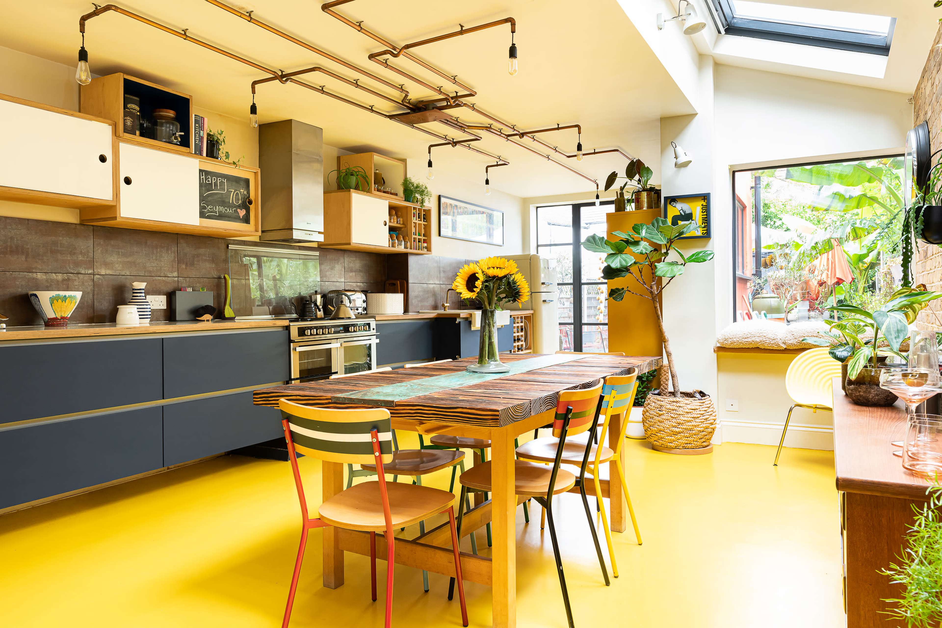 A modern kitchen with a large wooden dining table surrounded by colorful chairs, featuring yellow flooring and various plants in the space.
