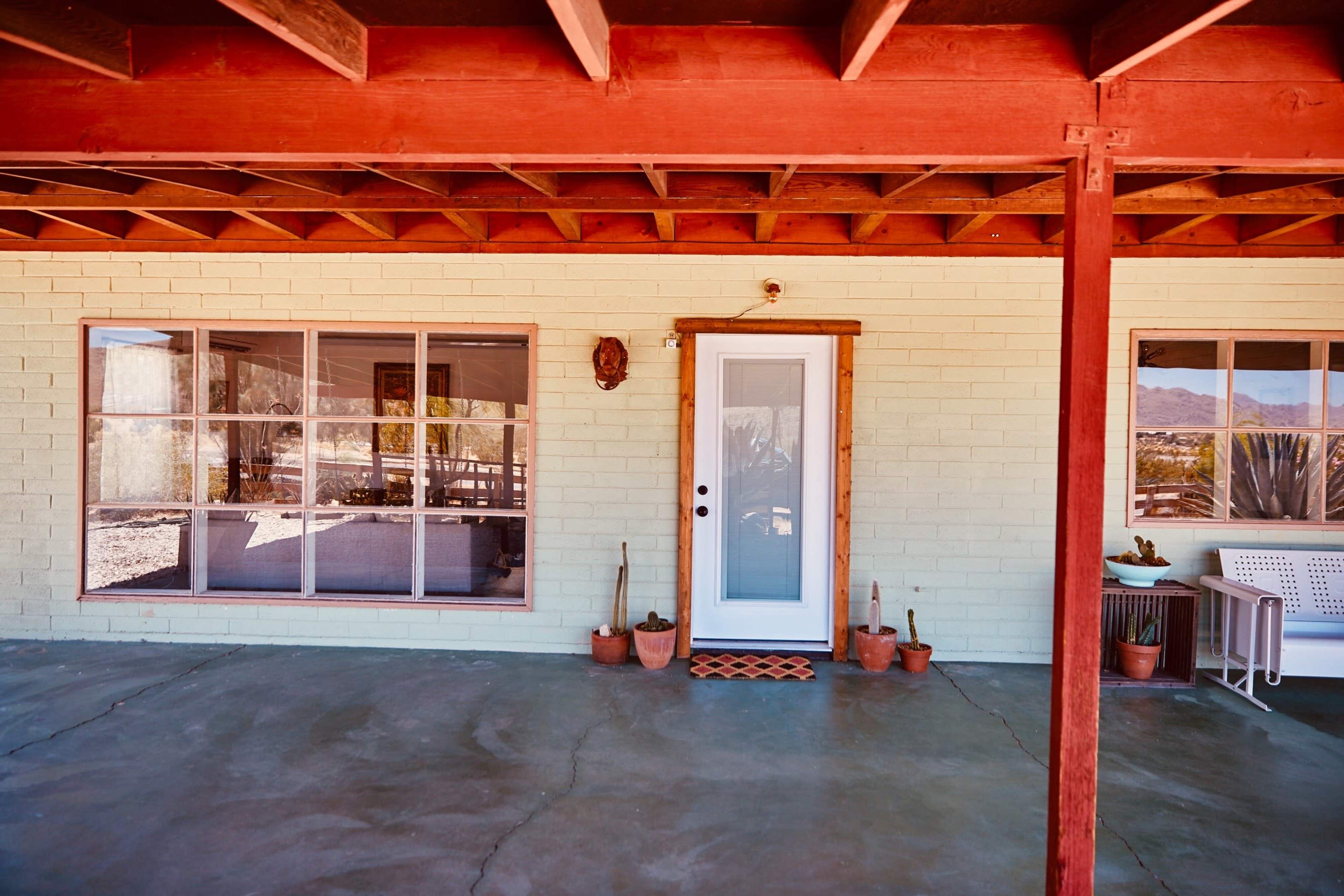 The image shows a front porch area of a building with a light-colored brick wall, a central door, large windows on one side, and potted plants.