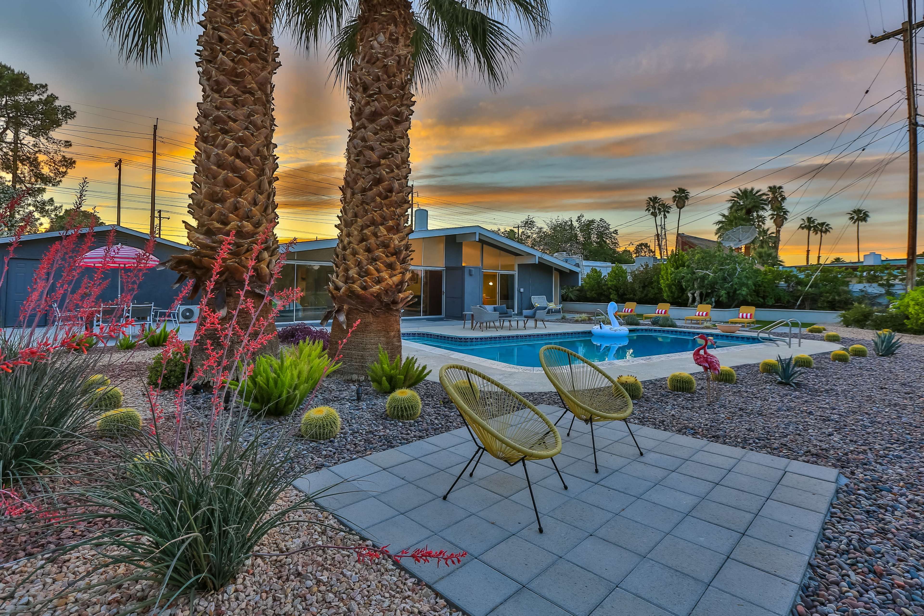 A landscaped backyard with a swimming pool, seating area, and palm trees under a colorful sunset sky.
