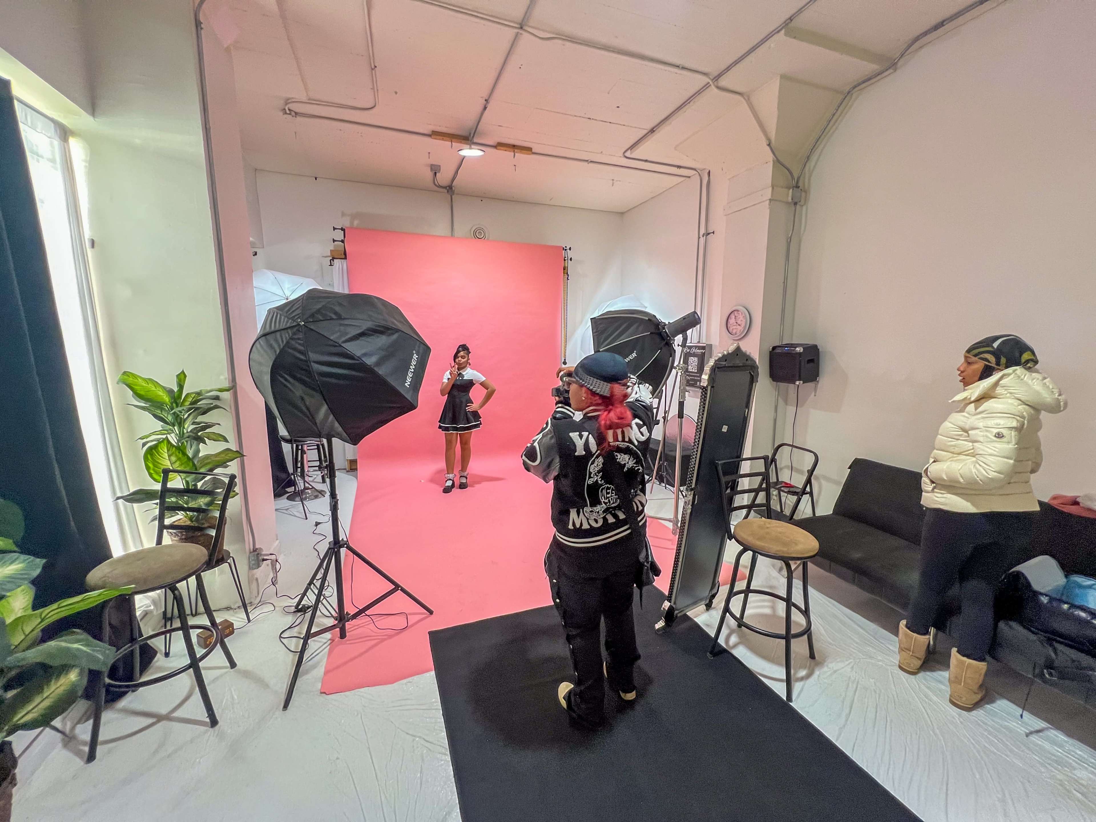 A photoshoot is taking place in a studio with a model posing in front of a pink backdrop while a photographer and another person are present.