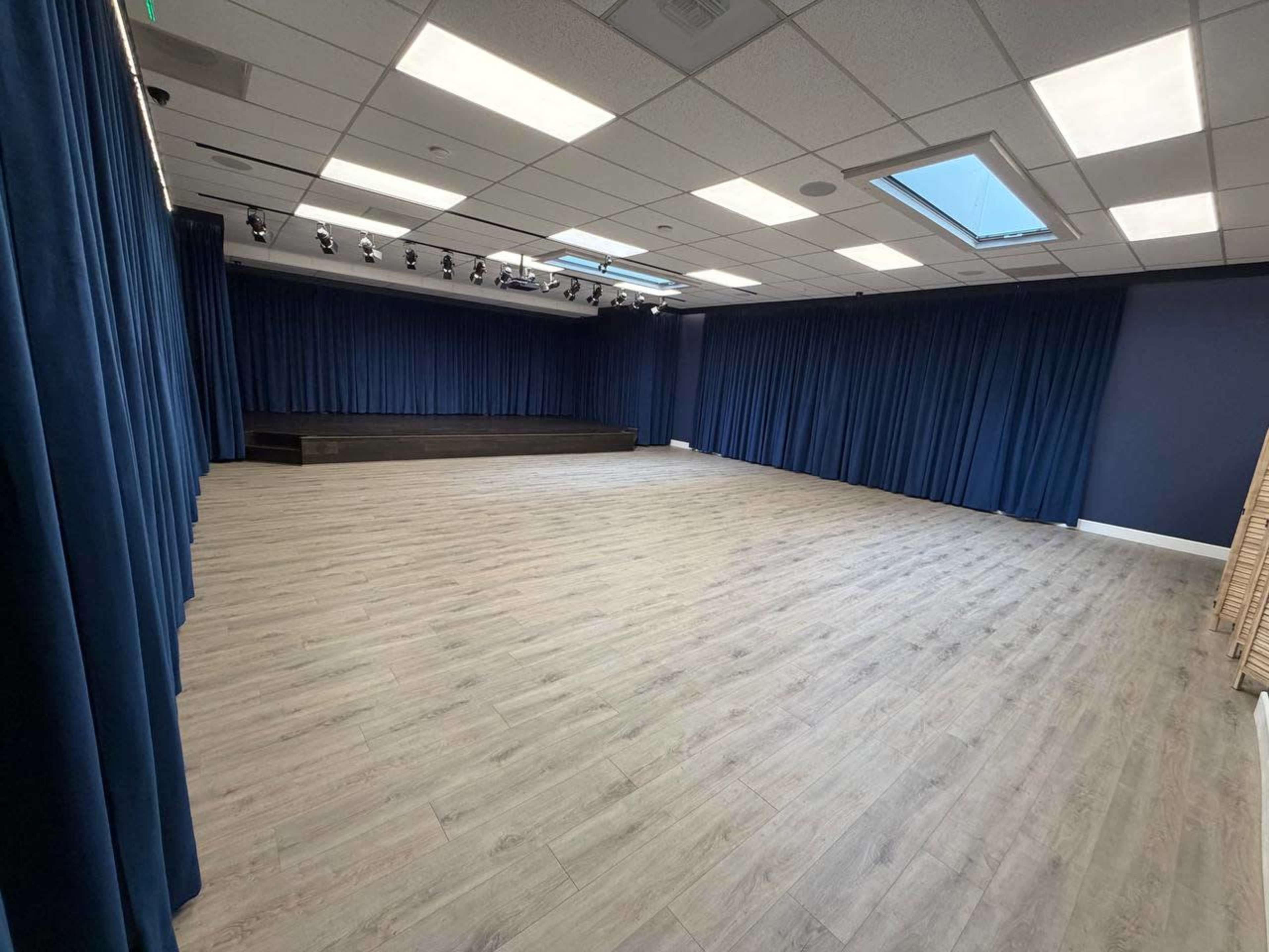 The image shows a spacious room with wooden floors, blue curtains lining the walls, and a raised platform at one end, illuminated by spotlights.