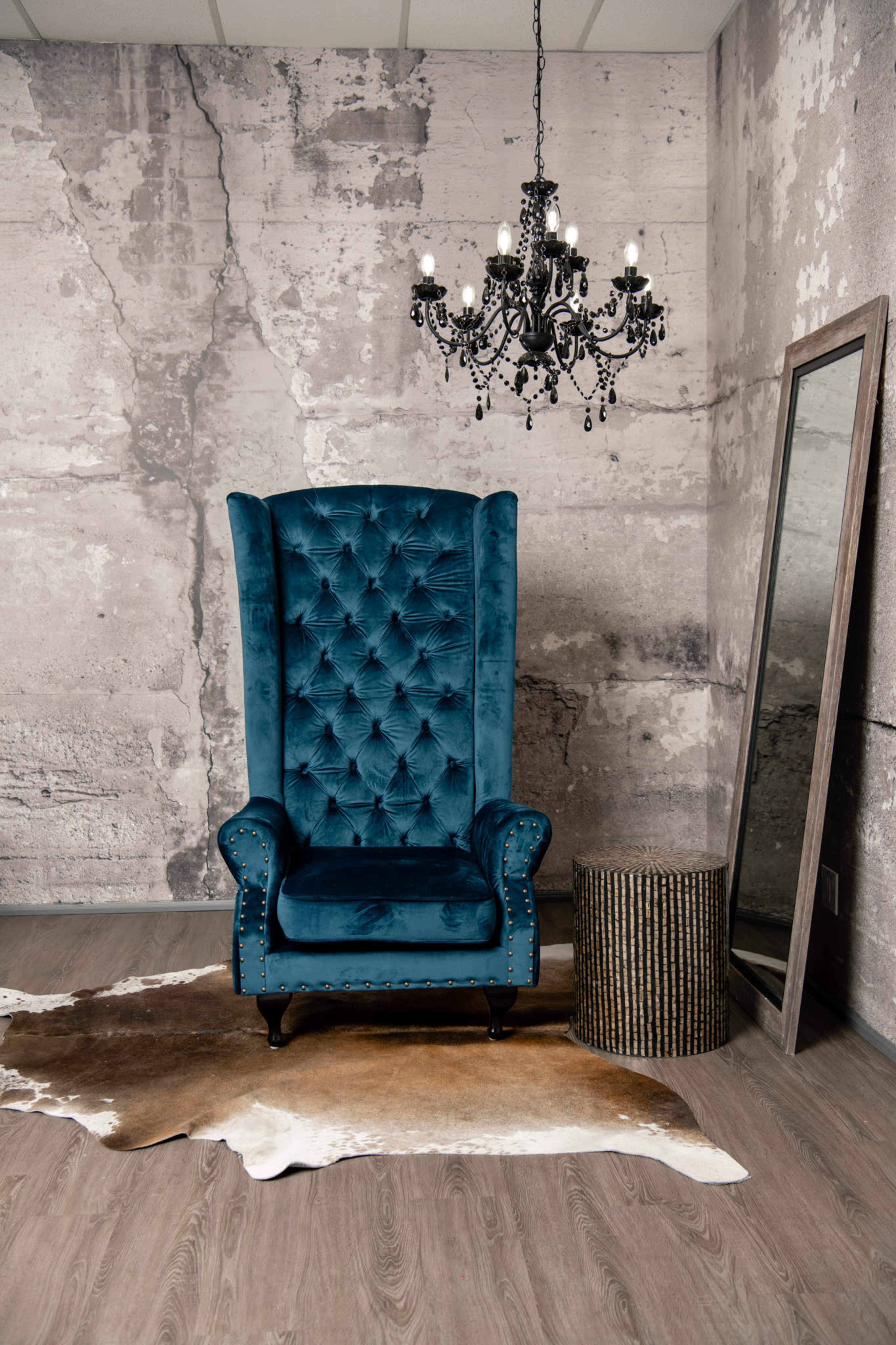 The scene features a blue velvet wingback chair beside a round, textured side table on a cowhide rug, with a vintage chandelier hanging overhead and a full-length mirror against a textured wall.