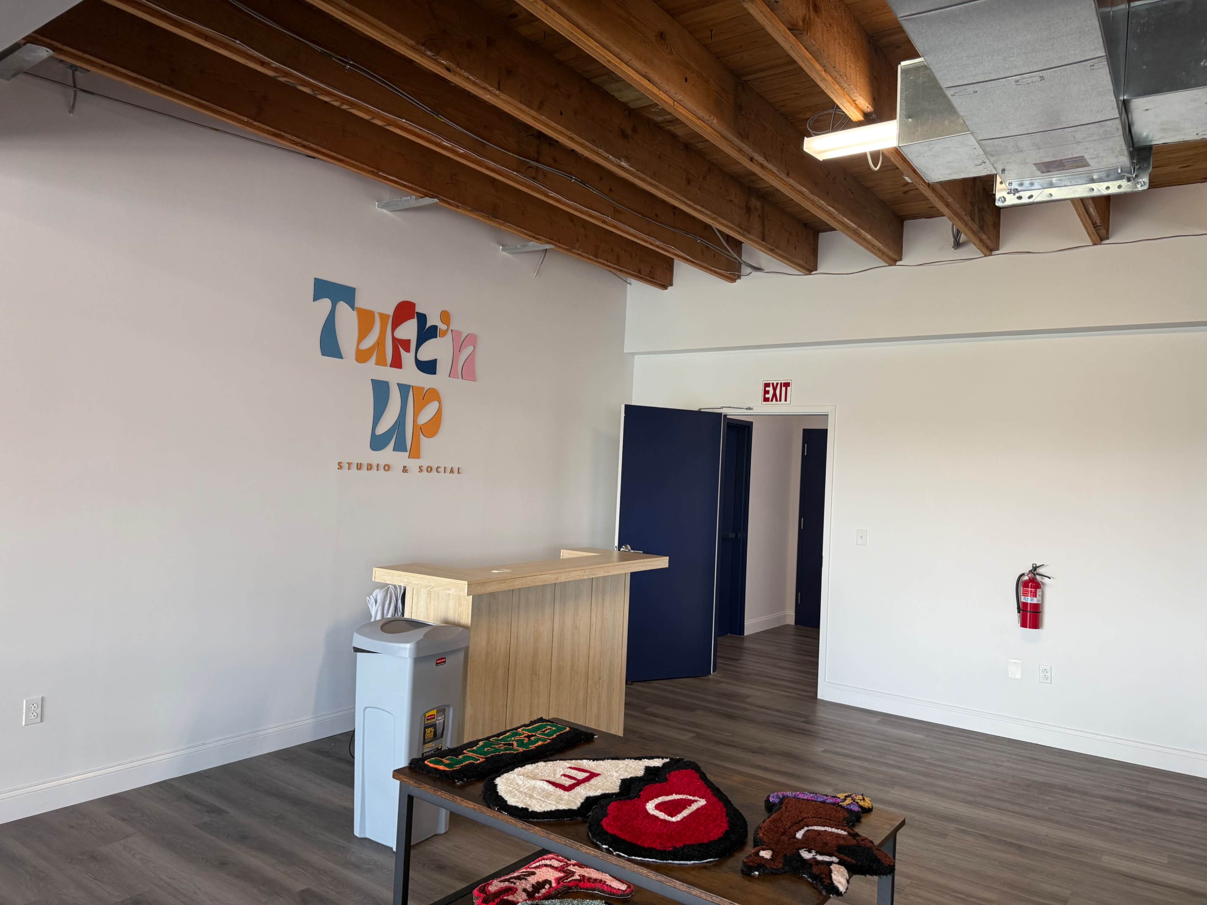 The image shows the interior of a modern studio space featuring a reception desk, a colorful wall sign that reads "Tuf’n Up Studio & Social," and hardwood ceilings.