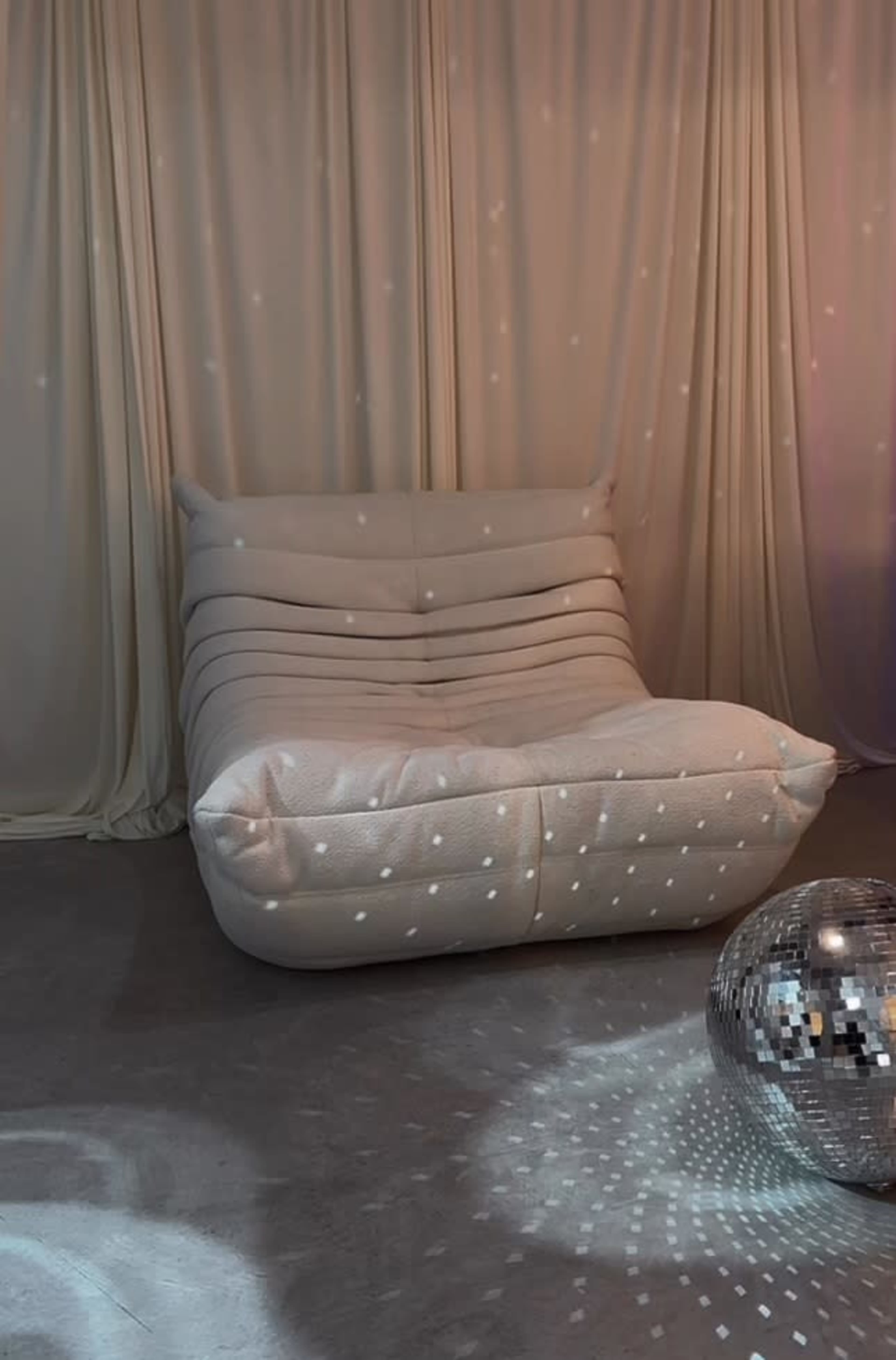 A textured white lounge chair sits against a backdrop of draped fabric, accompanied by a mirrored disco ball reflecting soft light patterns on the floor.