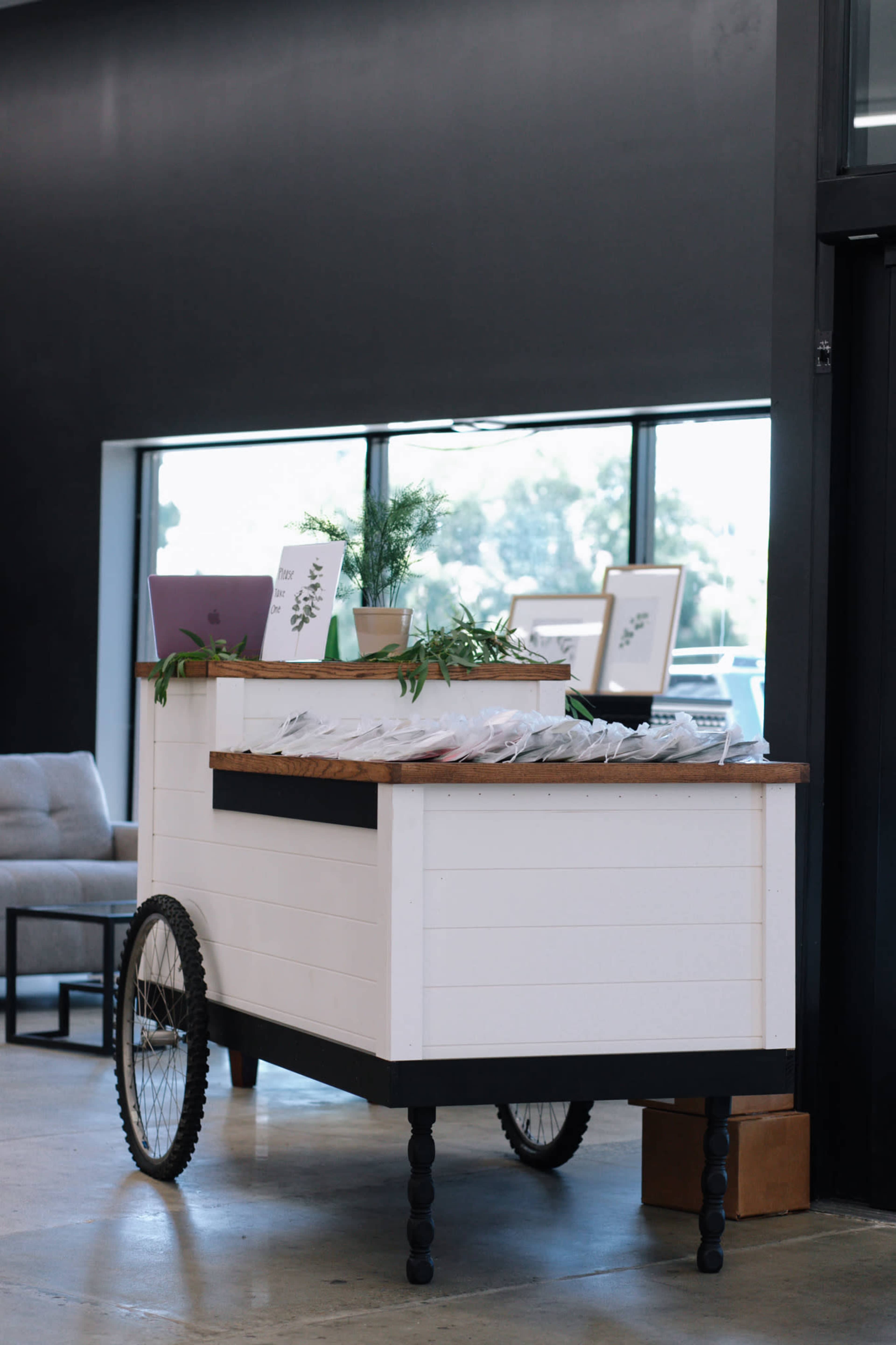 A white wooden cart with a wood top and black wheels stands beside a large window, displaying plants and framed items.