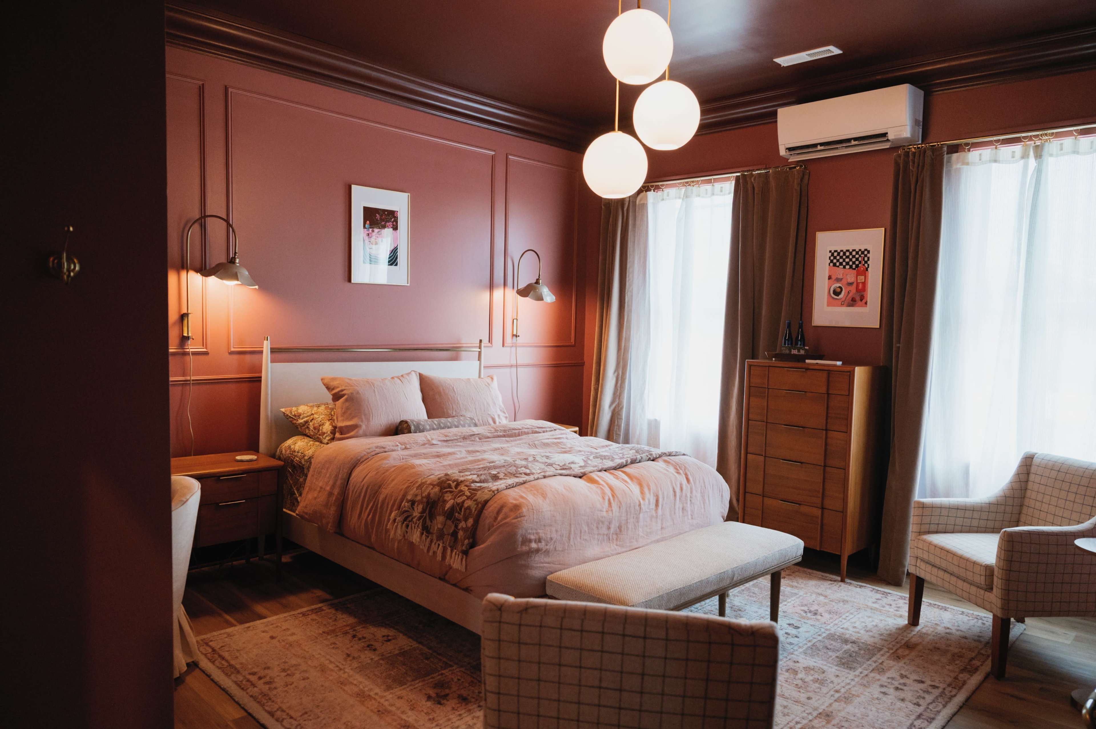 The image shows a stylish bedroom featuring a pink color scheme, a bed with decorative pillows, a wooden dresser, and large windows covered with curtains.