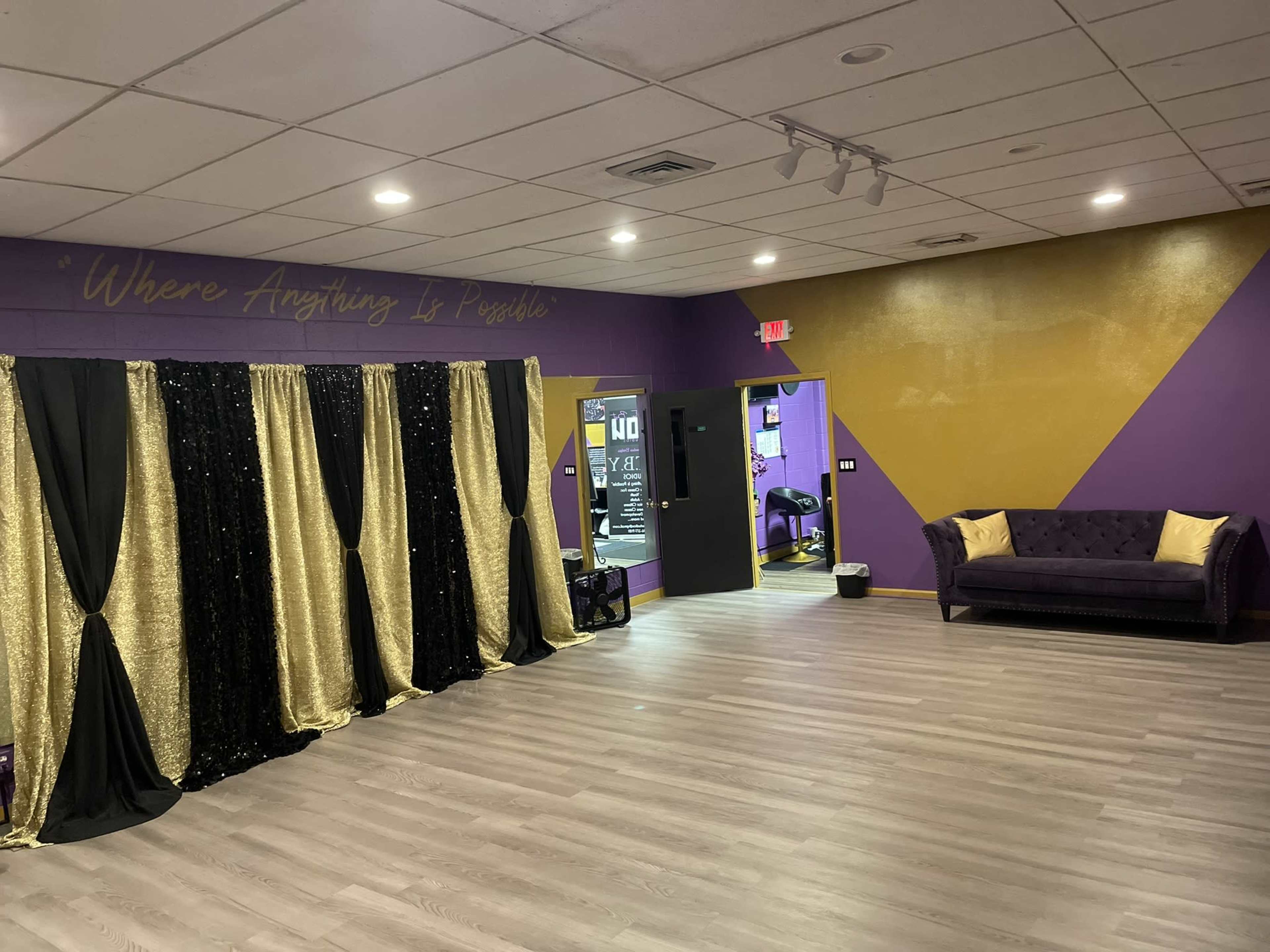 The image shows an interior space featuring a purple and gold color scheme, with a black and gold curtain setup on the left and a purple couch with cream-colored pillows on the right.
