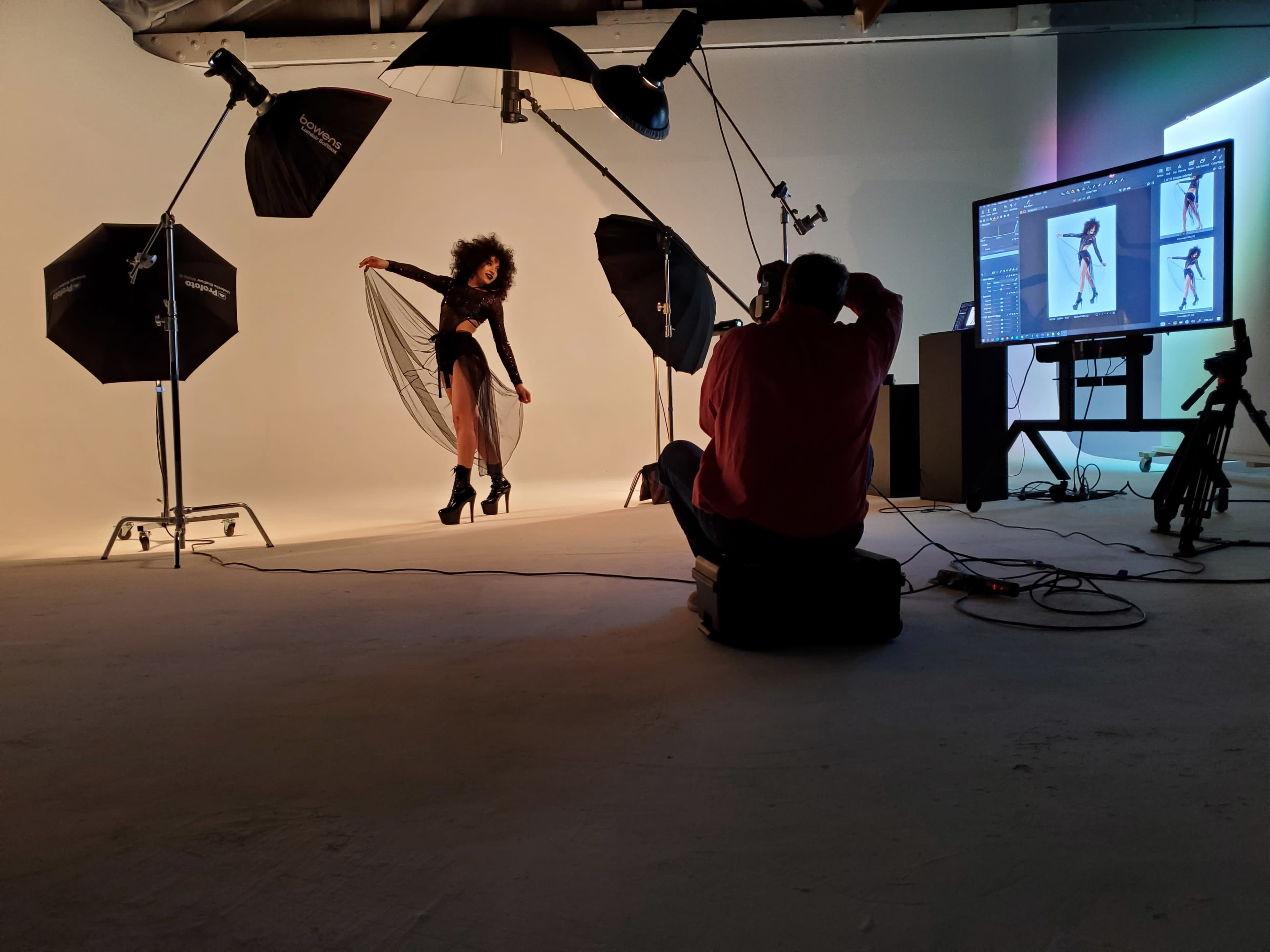 A model poses with a flowing dress on a photoshoot set, while a photographer reviews images on a computer screen.