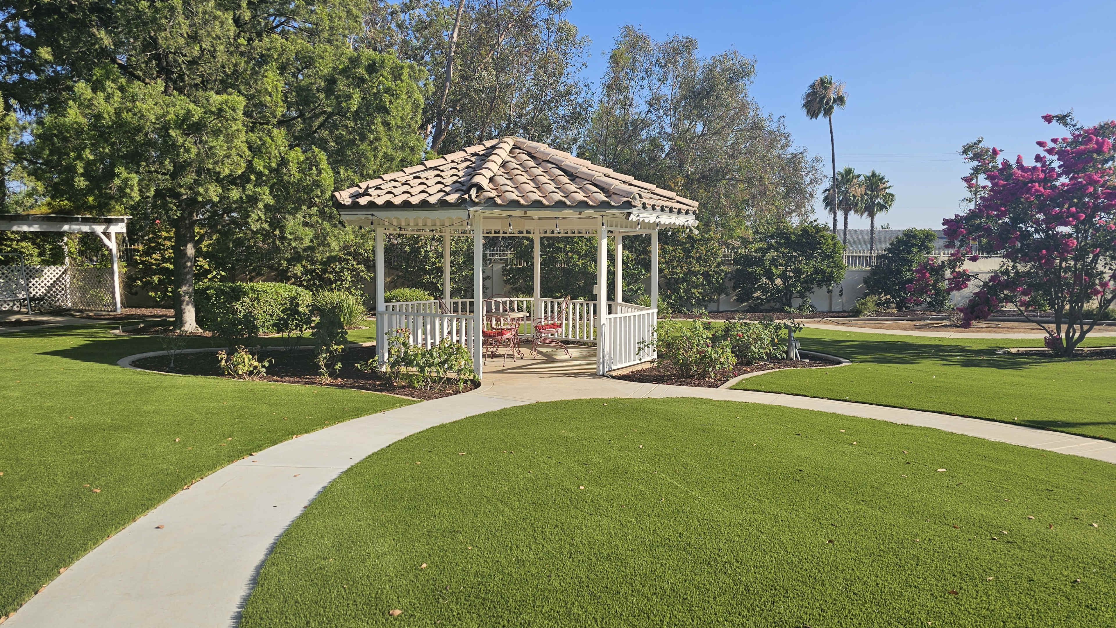 A white gazebo with a tiled roof is situated in a landscaped garden featuring green grass and flowering shrubs.