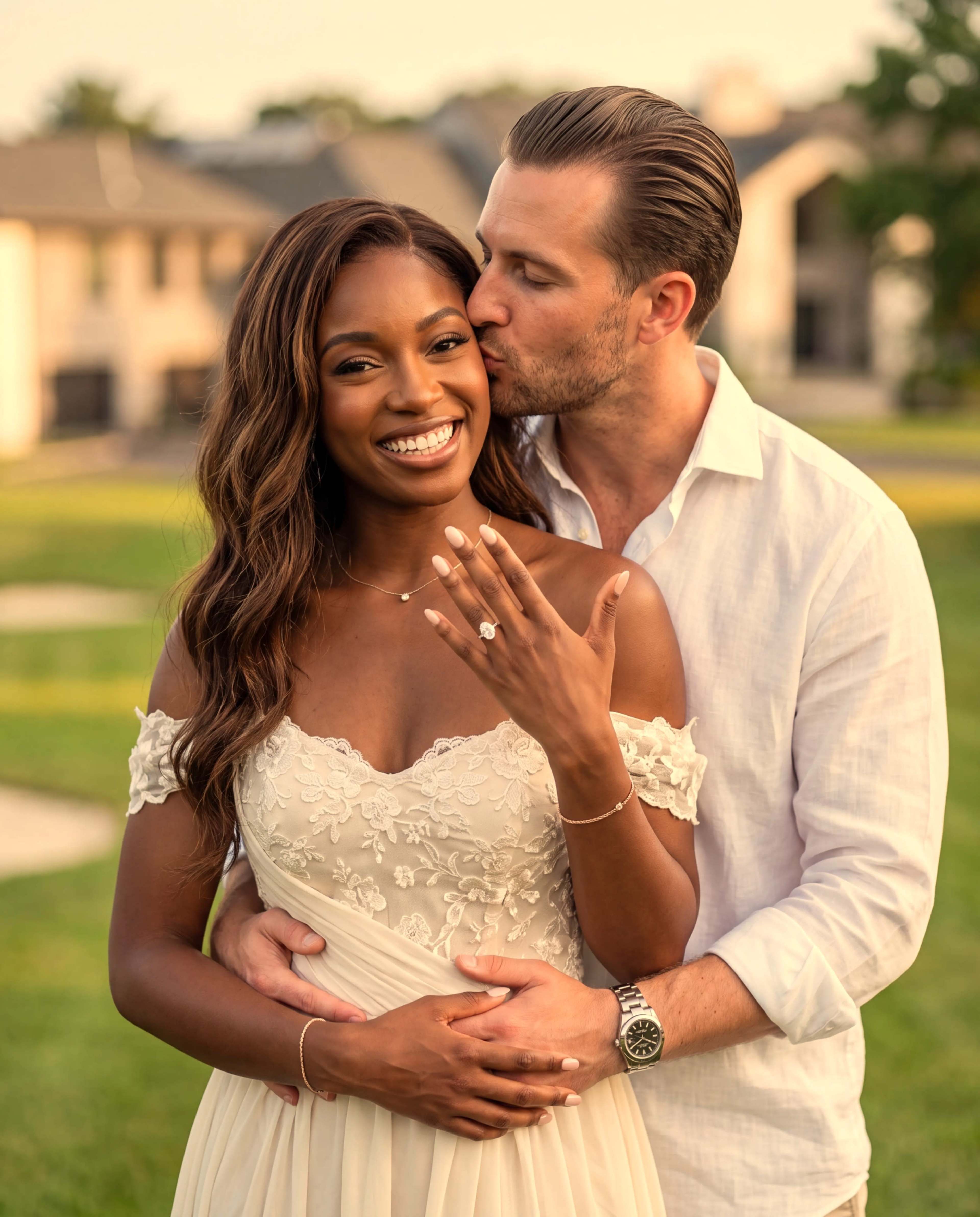 A couple stands outdoors, with the woman showing her engagement ring while the man kisses her on the cheek.