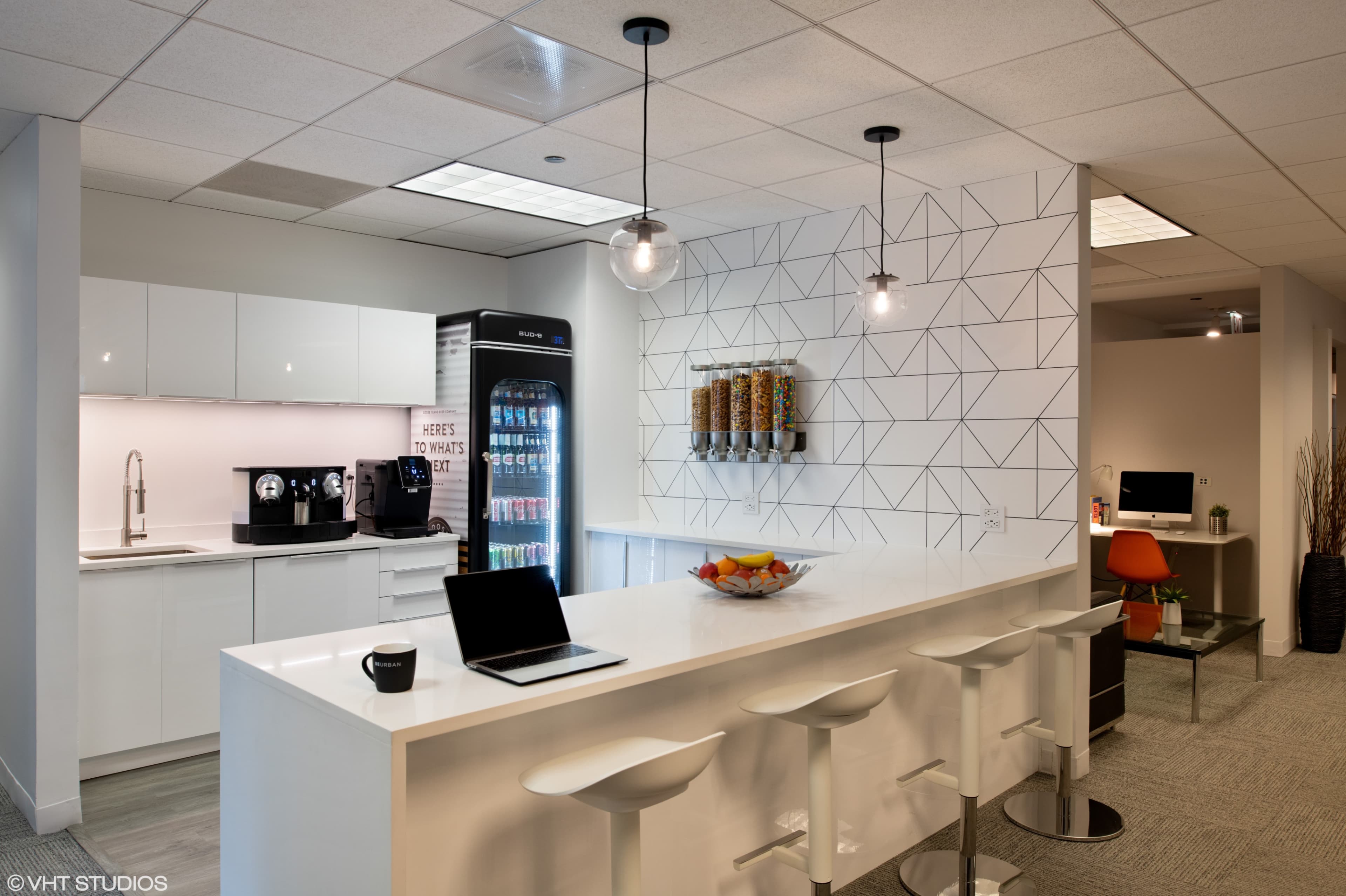 A modern office break room with a white counter, bar stools, a coffee machine, a vending machine, and a geometric-patterned wall.
