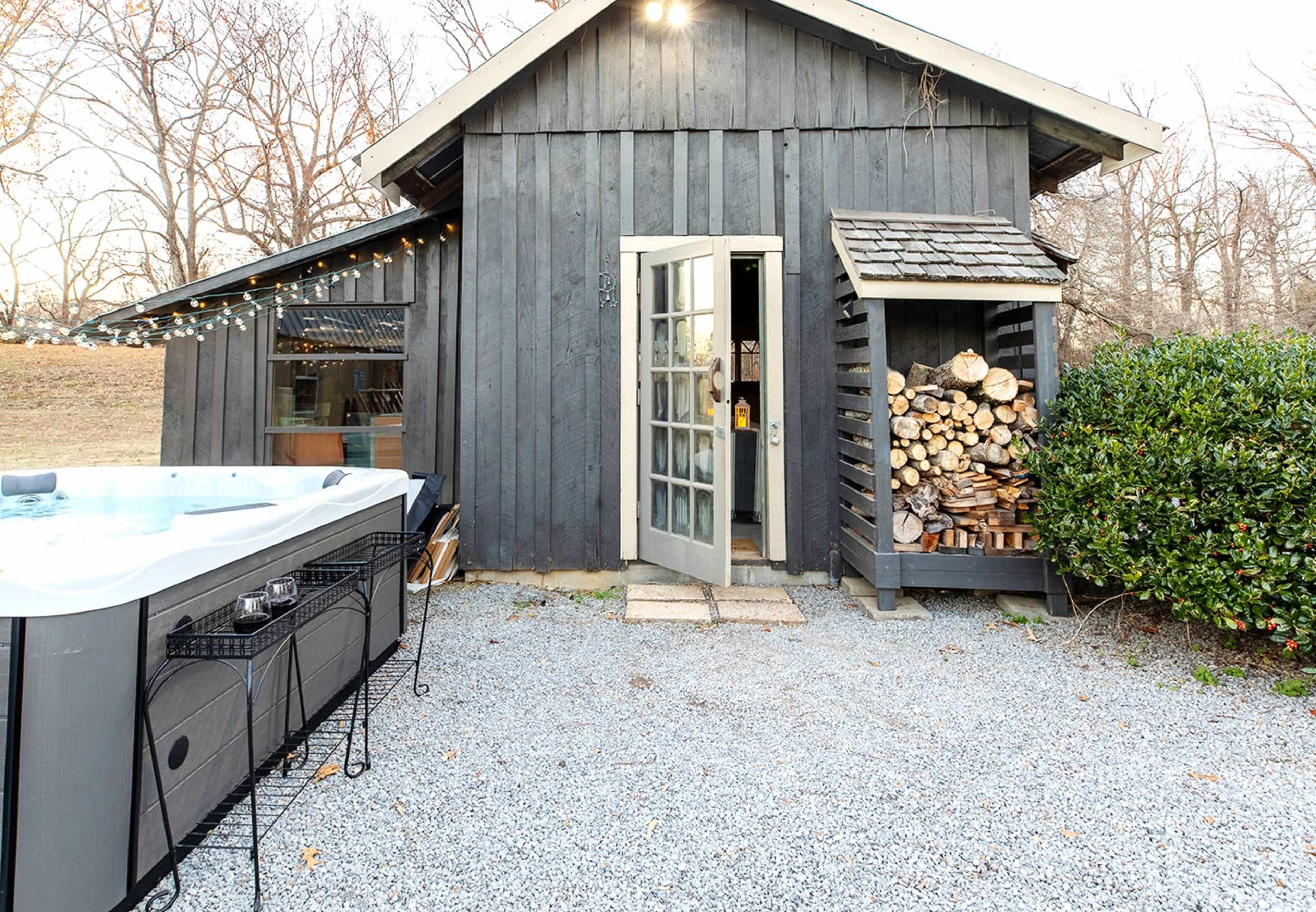 A gray wooden cabin with a hot tub on the left and a stack of firewood against the right side.