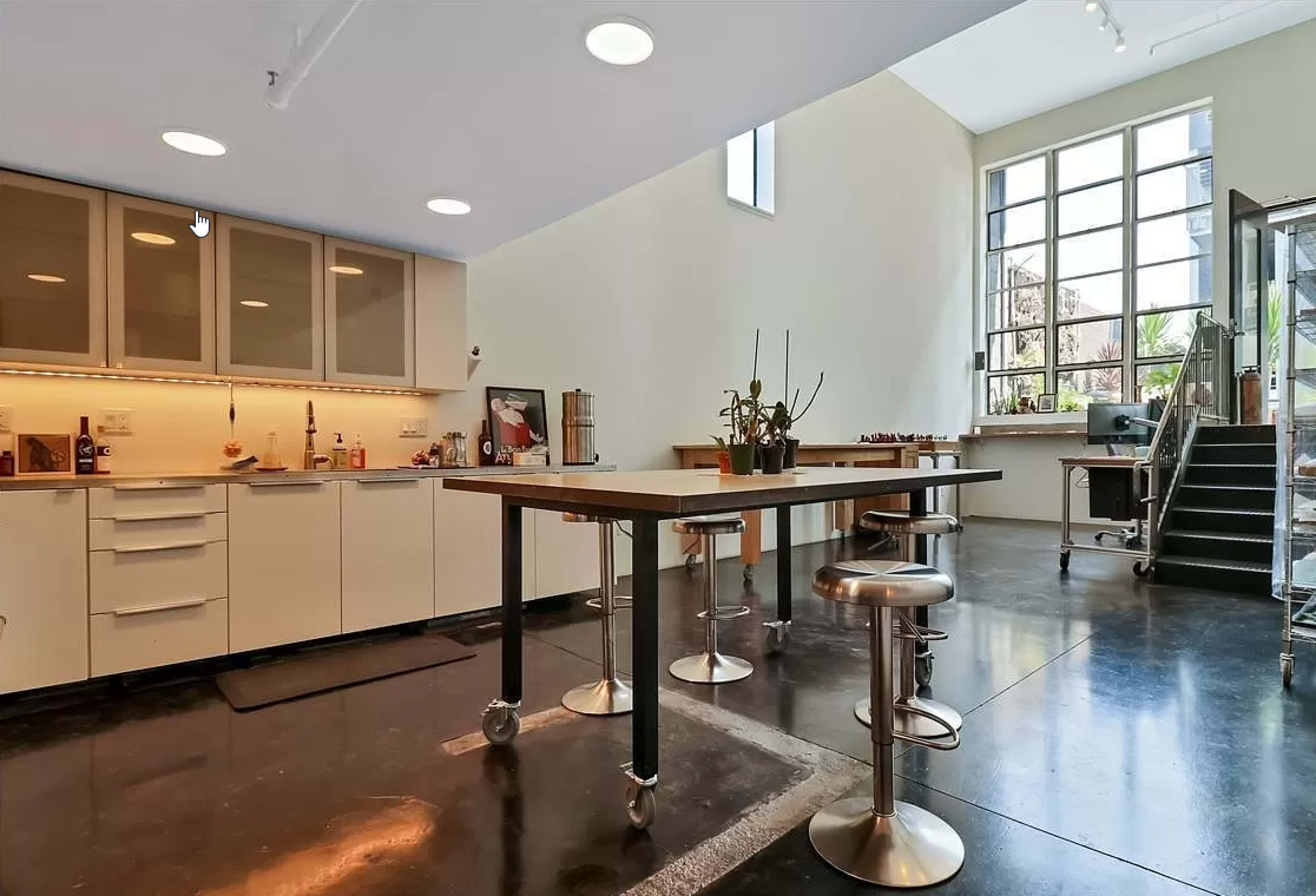 A modern kitchen with a central island and bar stools, featuring large windows and a staircase leading to an upper level.