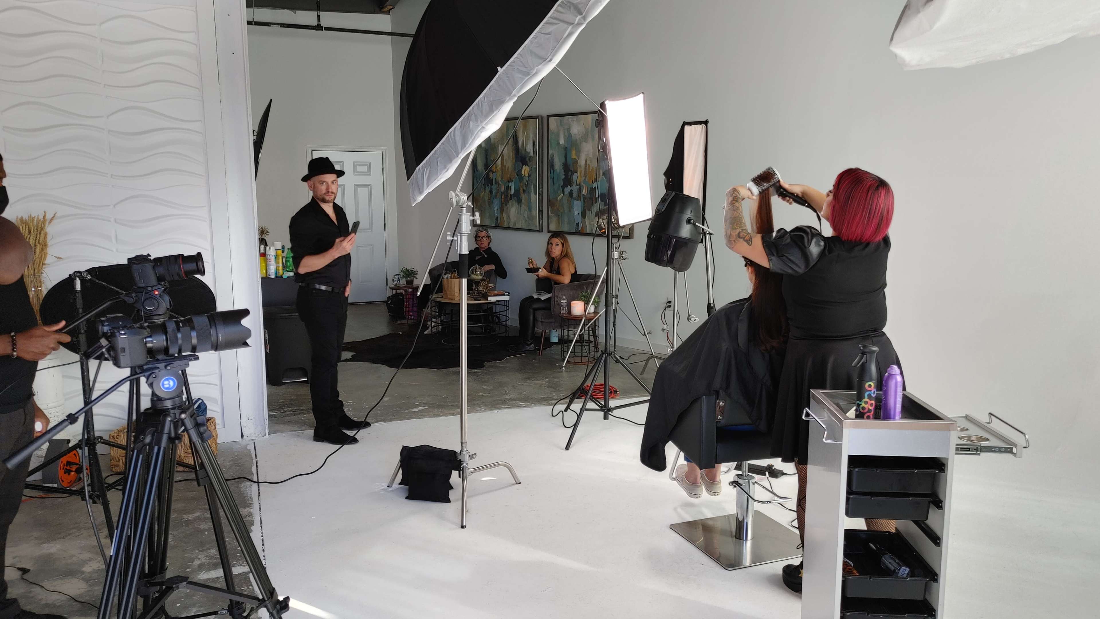 A hair stylist works on a client's hair in a studio setting, while a photographer and two models observe in the background.