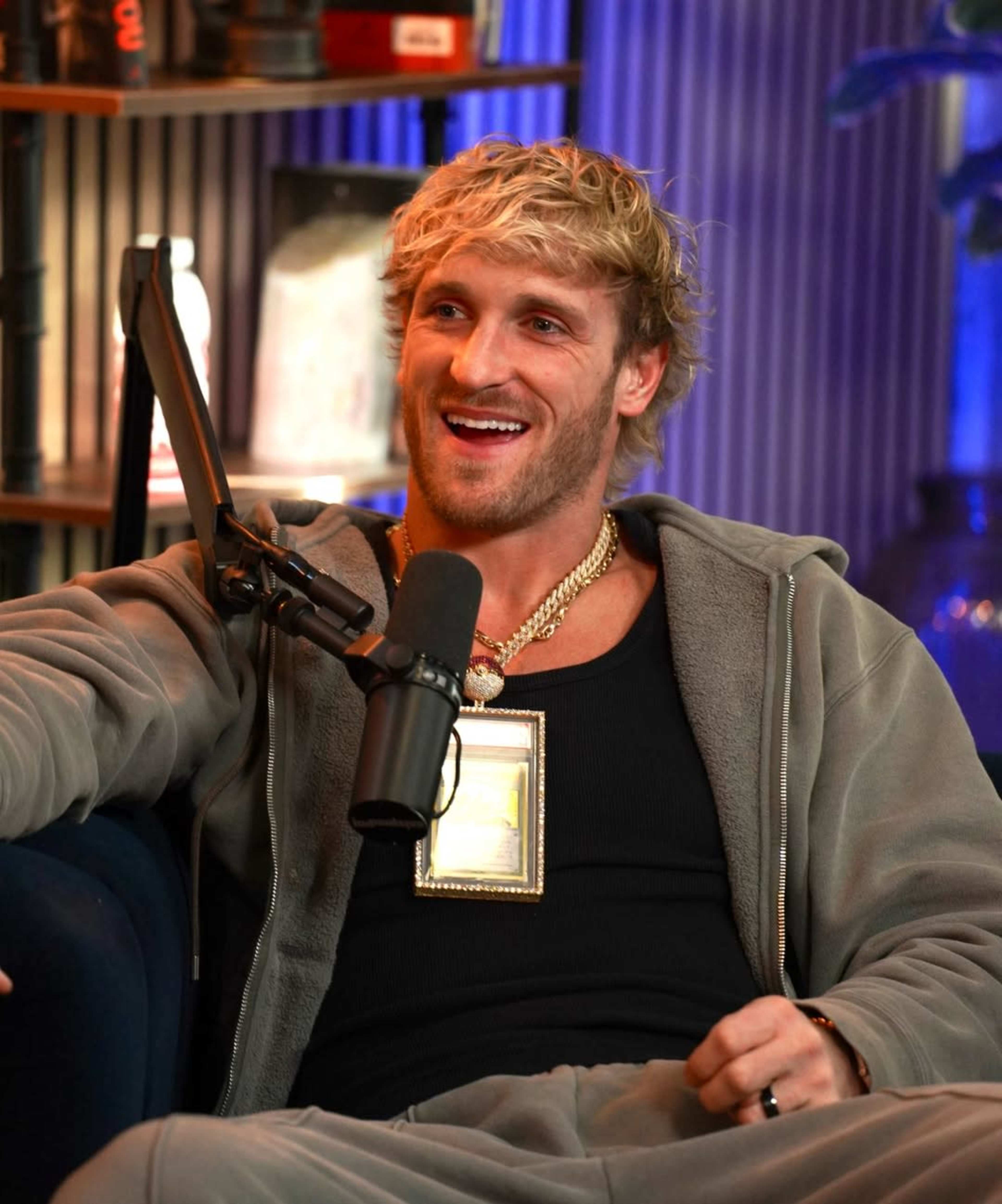 A man with curly blonde hair is sitting on a blue couch, wearing a gray tracksuit and a large pendant around his neck, while speaking into a microphone.