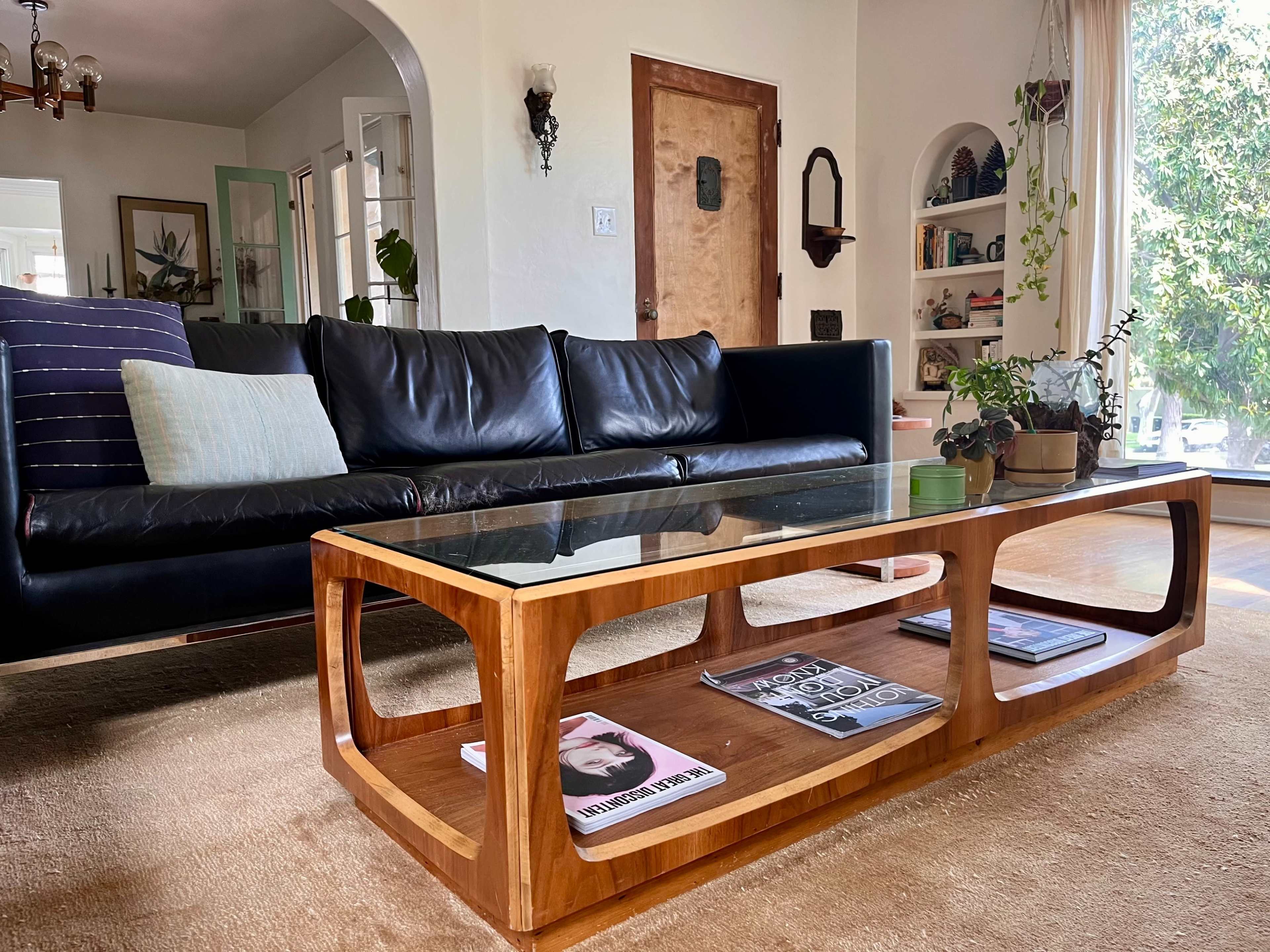 The image shows a modern living room with a black leather sofa, a wooden coffee table featuring magazines underneath a glass top, and potted plants in a well-lit space.